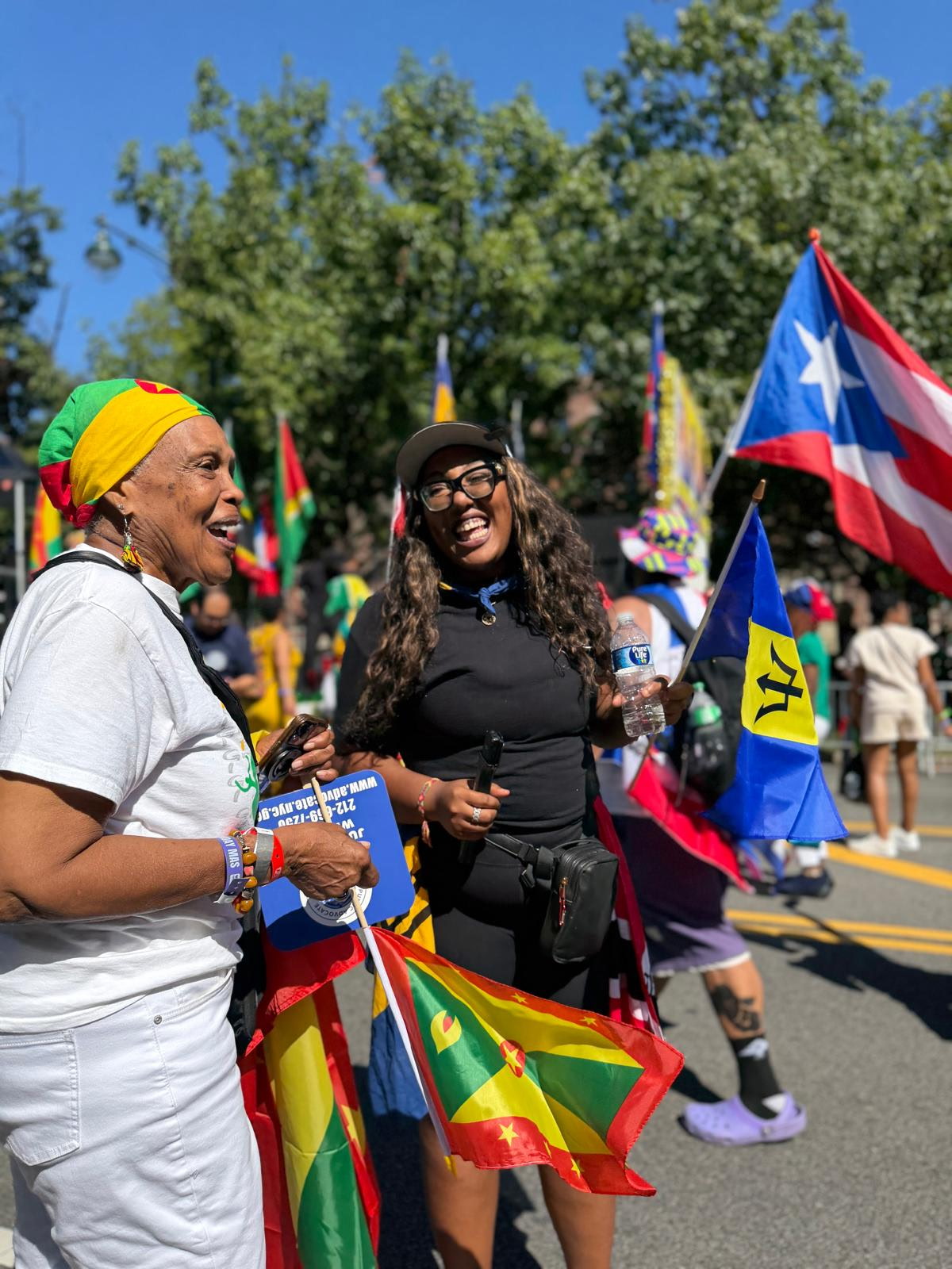 Two women smiling at a parade with flags and trees in the background.