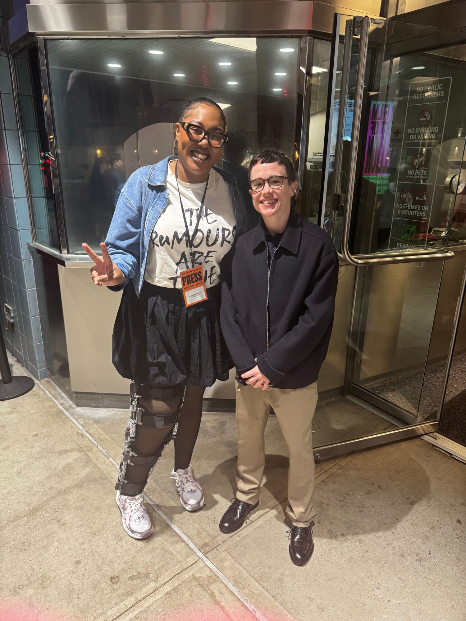Two people standing in front of a glass door, one wearing a black jacket and the other in a blue jacket, both smiling.