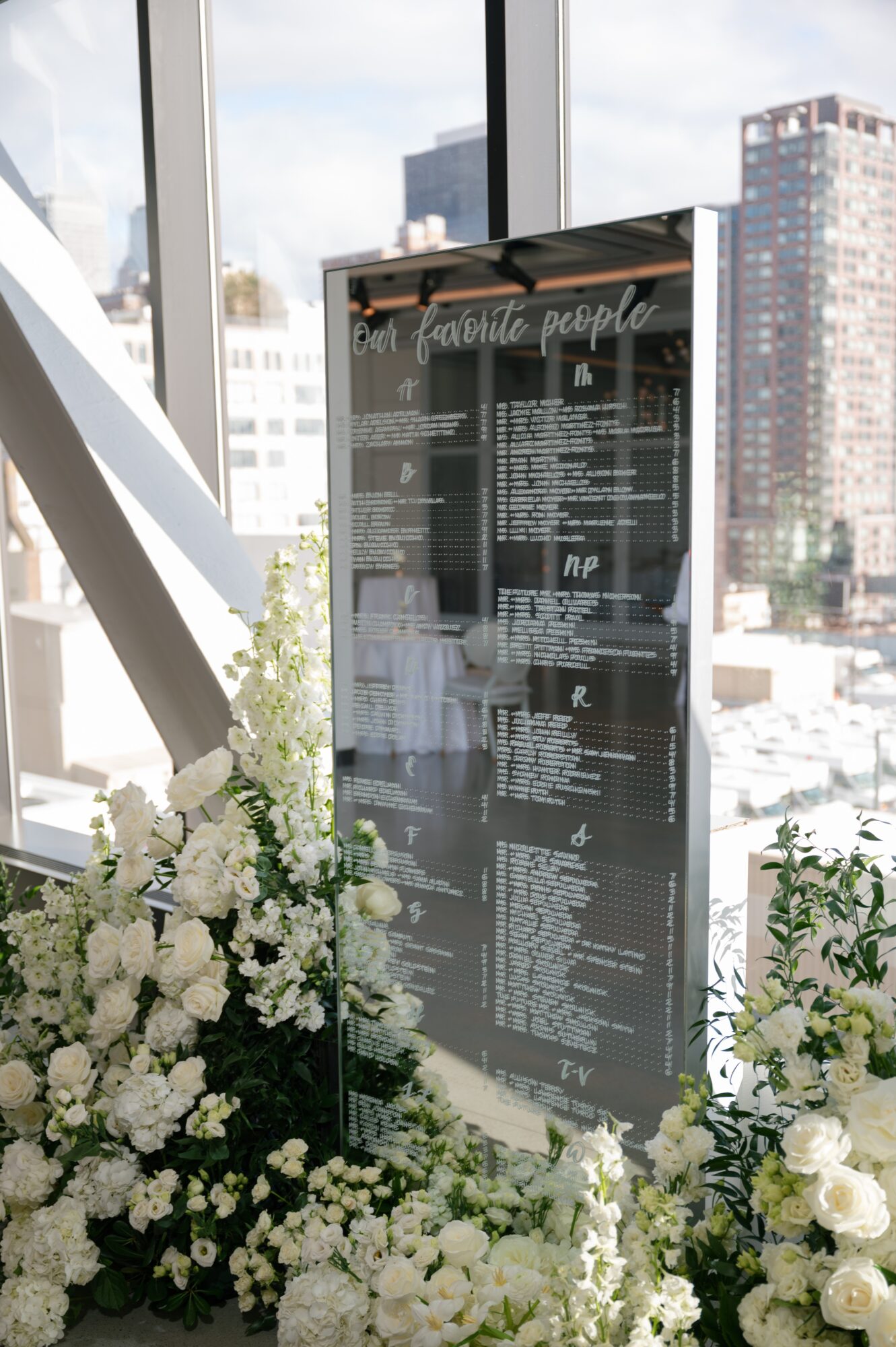 A tall, black menu board with white text stands near large windows with a cityscape view, surrounded by white flowers.