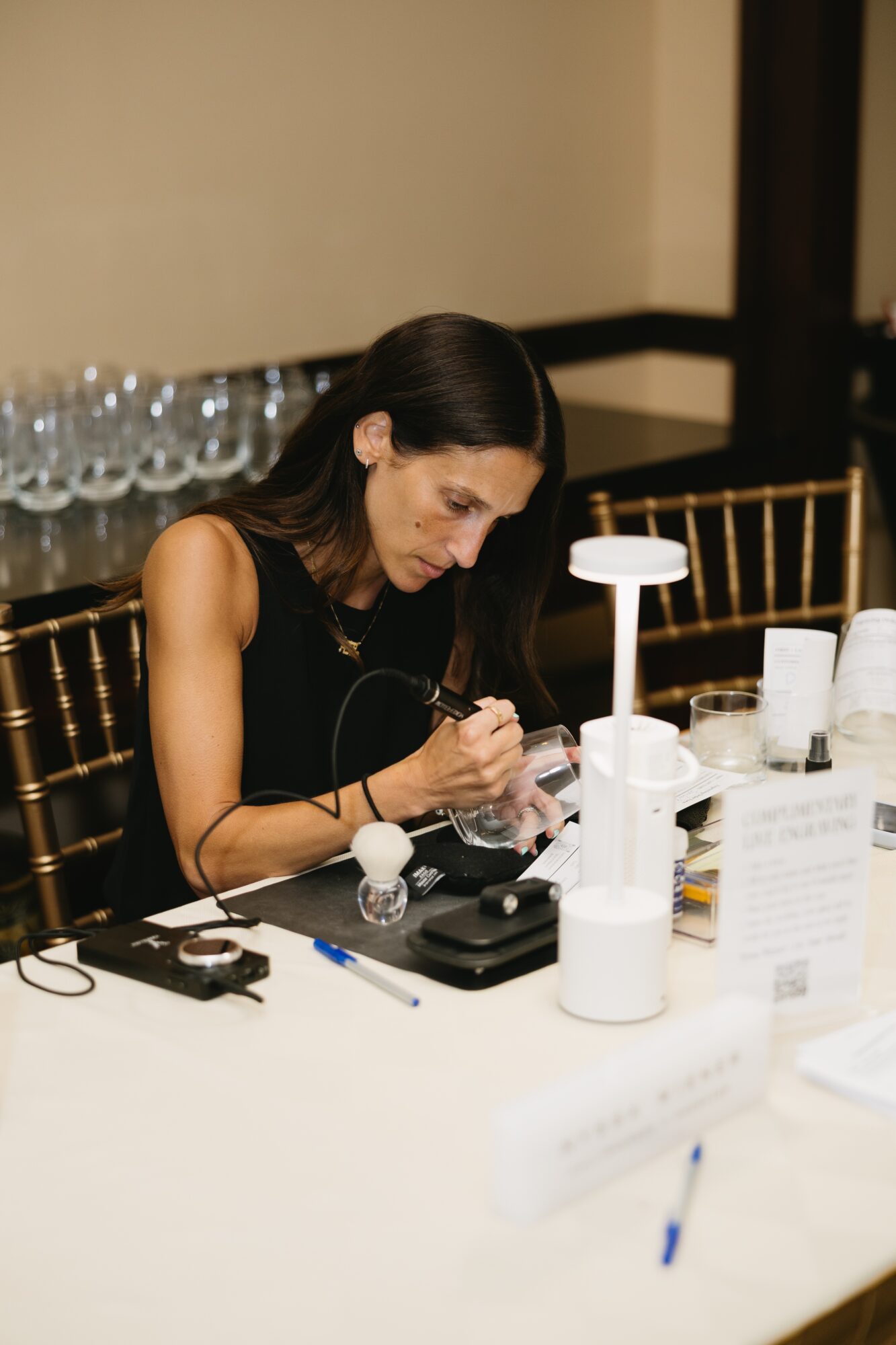 Woman with dark hair working at a table with various tools and equipment, focused on her task.