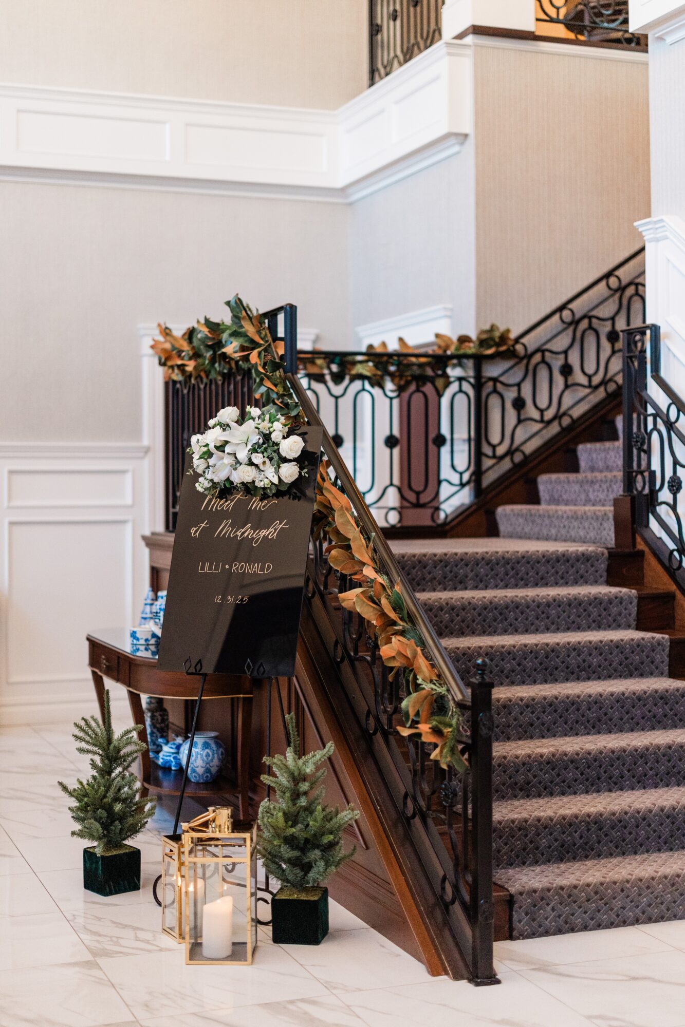 Decorated staircase with floral arrangements, a sign, and potted plants at the base, inside a building with white walls and a black railing.