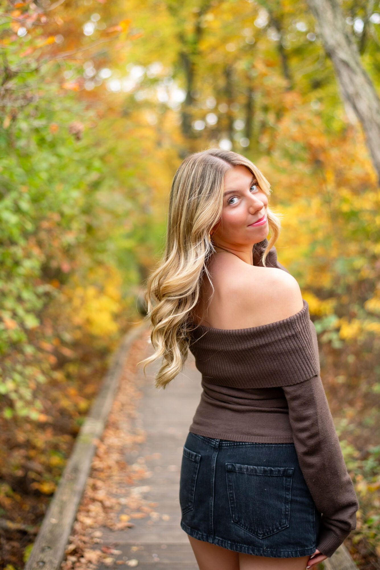 Young woman with long wavy hair standing on a forest path surrounded by autumn trees, looking over her shoulder.