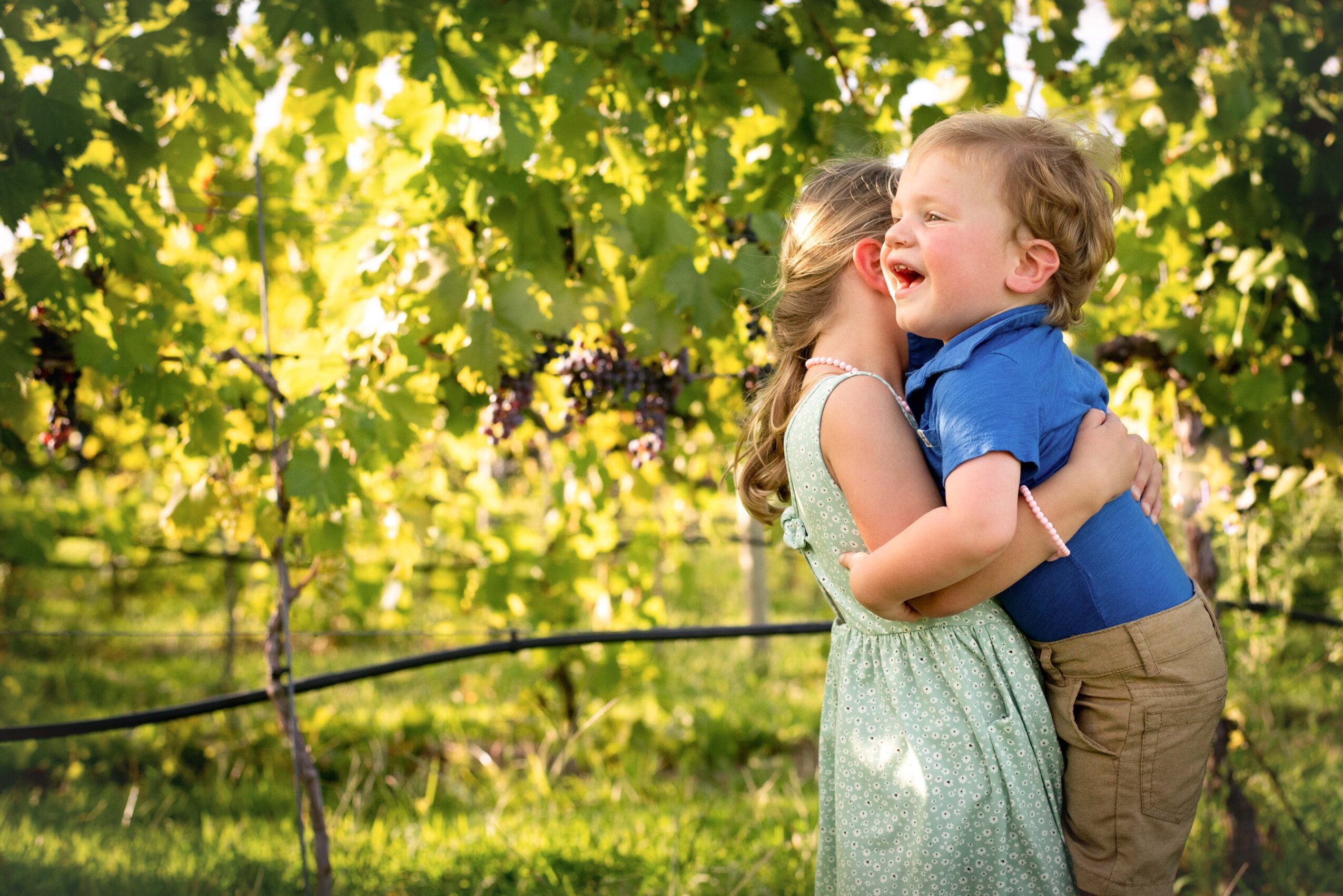 Two children, a girl and a boy, hugging and laughing outdoors with green trees and sunlight in the background.