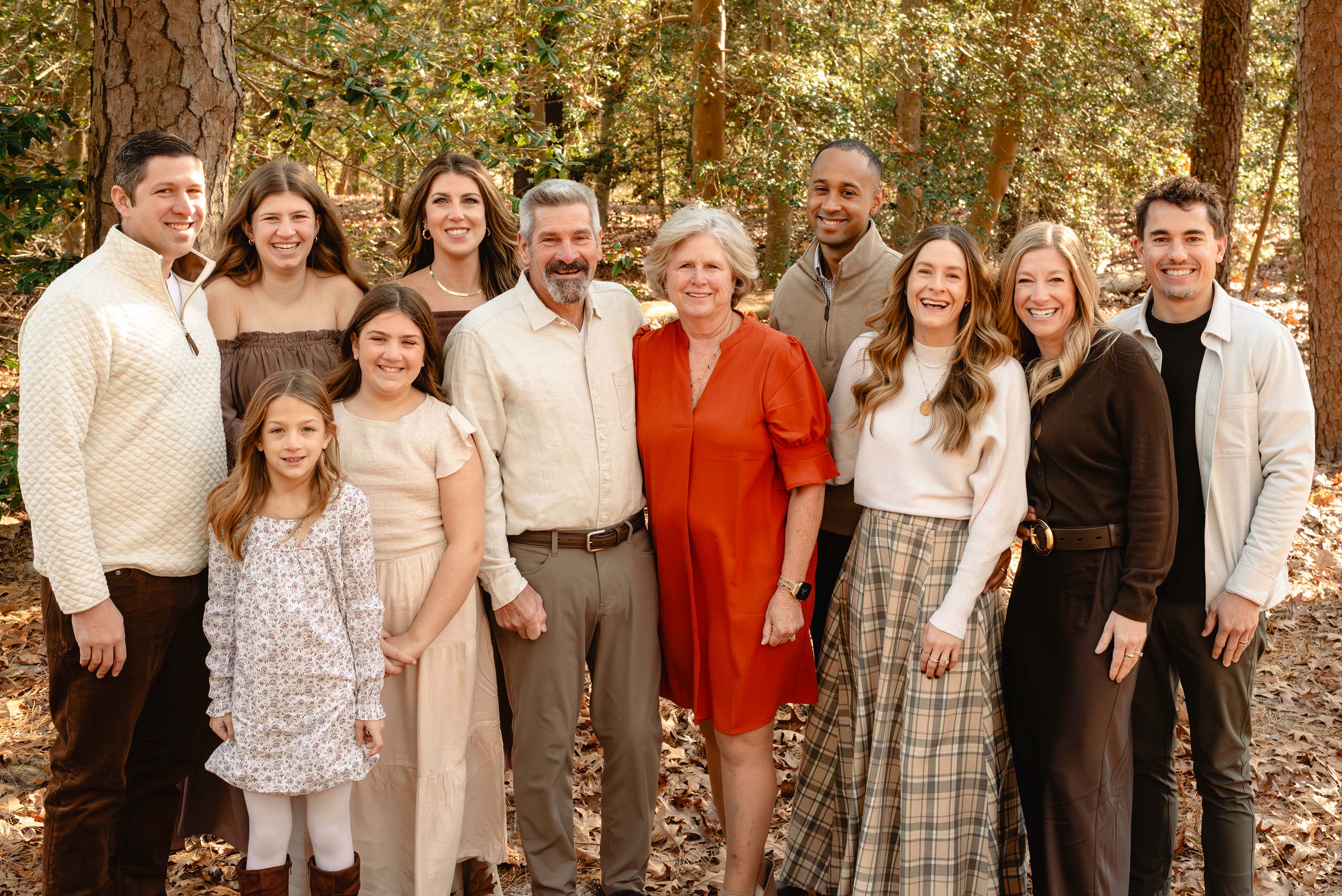 Group of ten people standing outdoors in a forest, smiling, with trees and sunlight in the background.