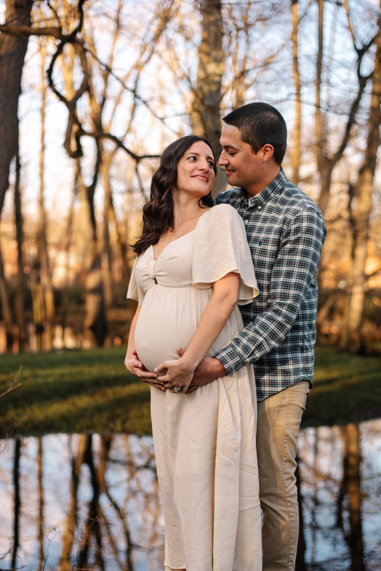 Pregnant woman and man standing outdoors near a pond, holding her belly, trees in background, sunset lighting.