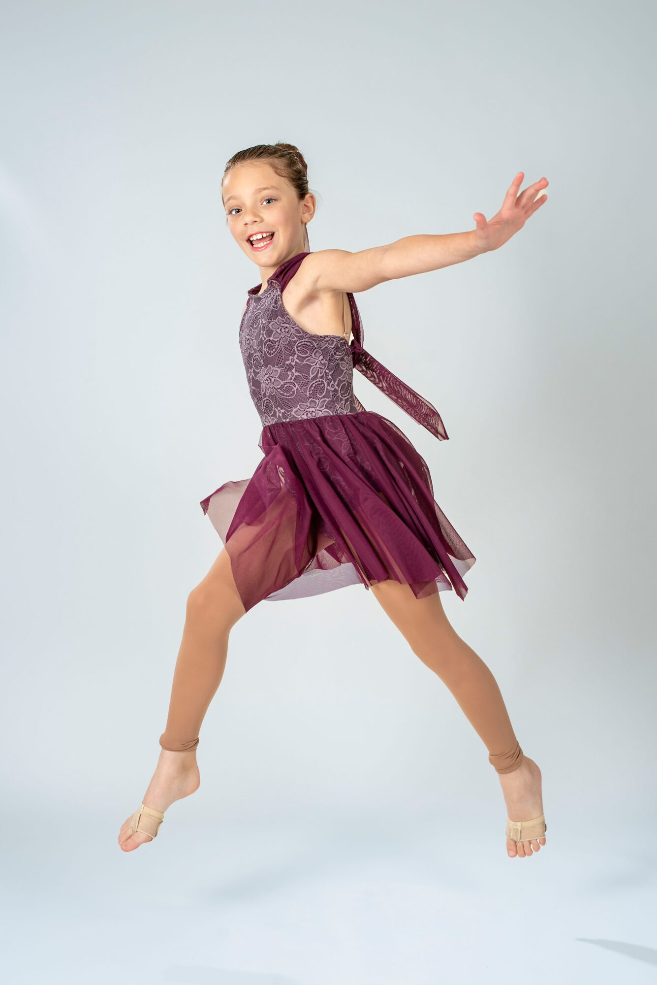 Young girl in a purple dress jumping with arms outstretched, smiling, against a light background.
