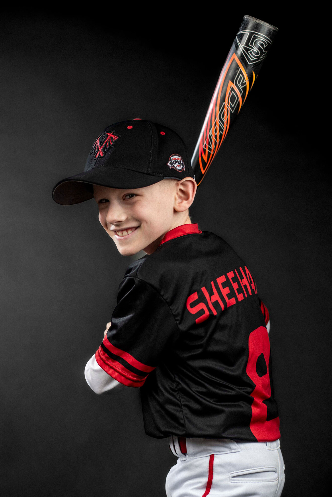 Young baseball player smiling, wearing a black cap and jersey, holding a bat over shoulder, dark background.