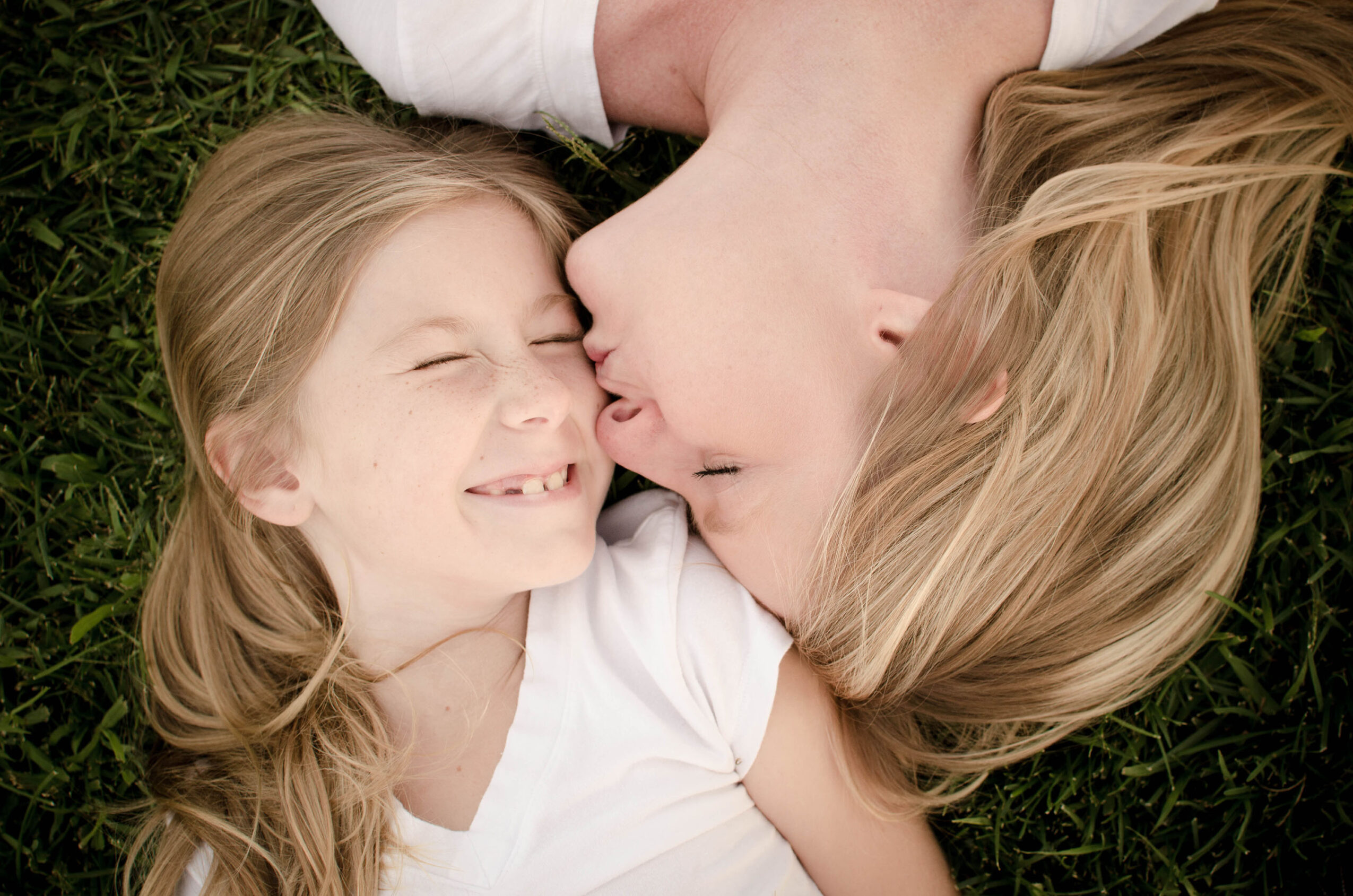 A woman and a girl lie on grass, face to face, smiling and about to kiss.