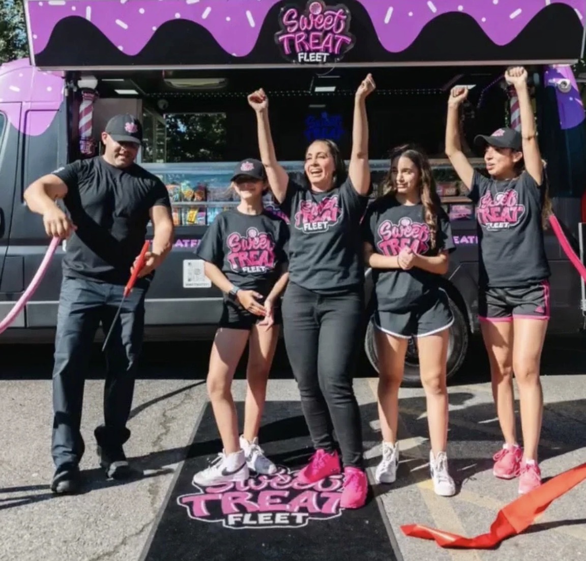 Group of five people celebrating in front of a food truck, some raising fists, wearing matching black shirts, pink shoes, and caps.