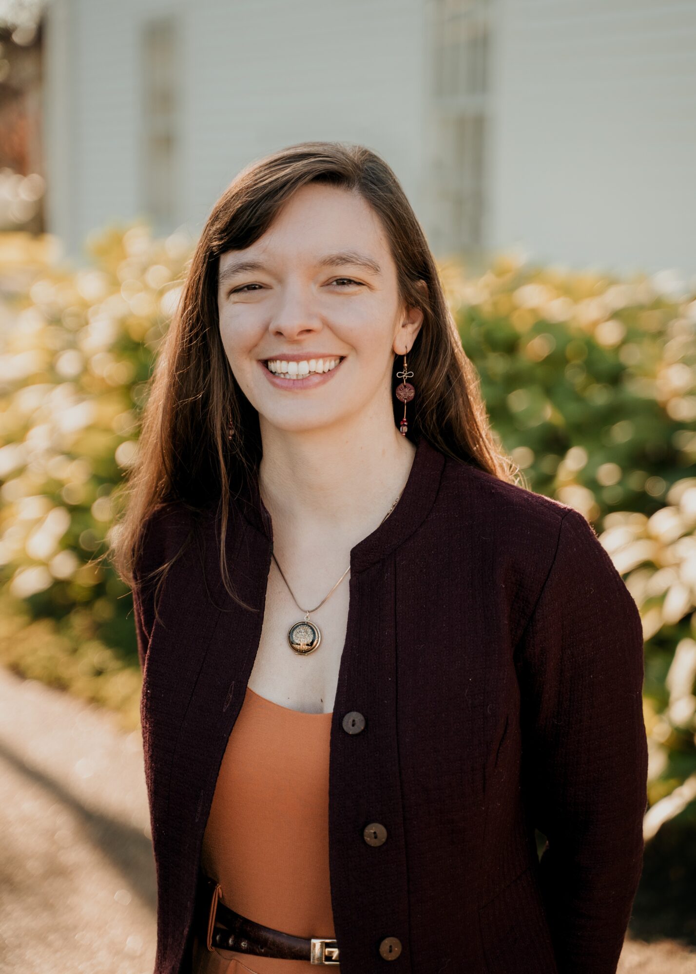 Young woman with long brown hair smiling outdoors, wearing a dark cardigan and orange top, with blurred background of plants and buildings.