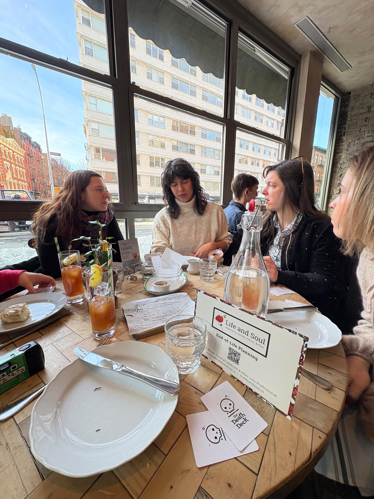 Group of women sitting at a table in a restaurant with large windows, drinks, and plates, engaging in conversation.