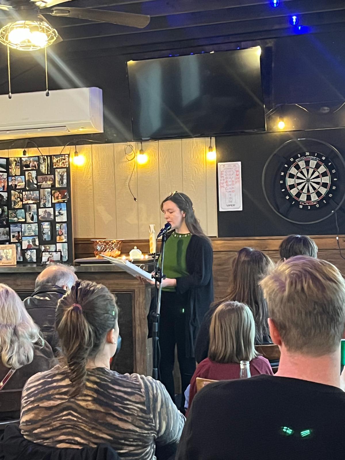 Woman singing into microphone in a bar with audience, dartboard, and TV screen behind her.