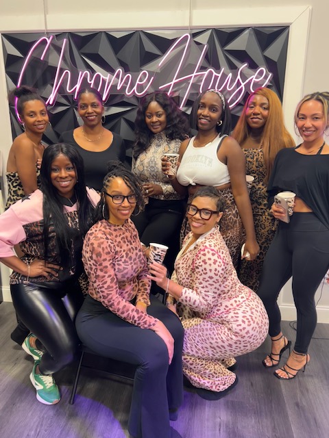 Group of ten women smiling, some holding drinks, in front of a neon sign that says 'Chyme House'.