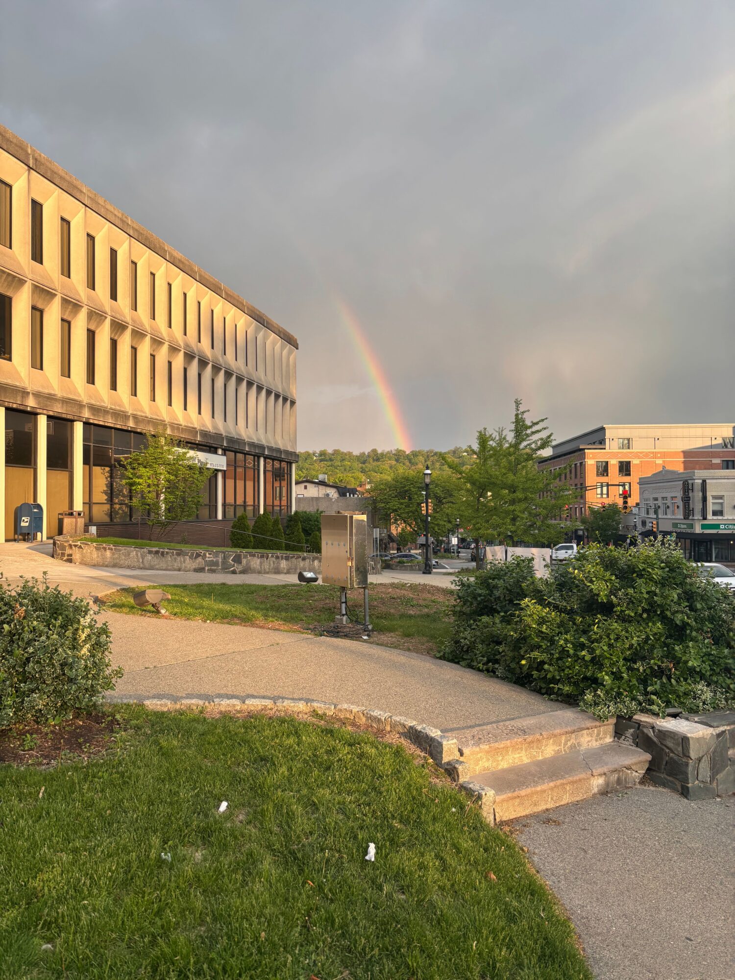 Street scene with a rainbow, buildings, trees, and a sidewalk, viewed from the side with a curved grassy area.
