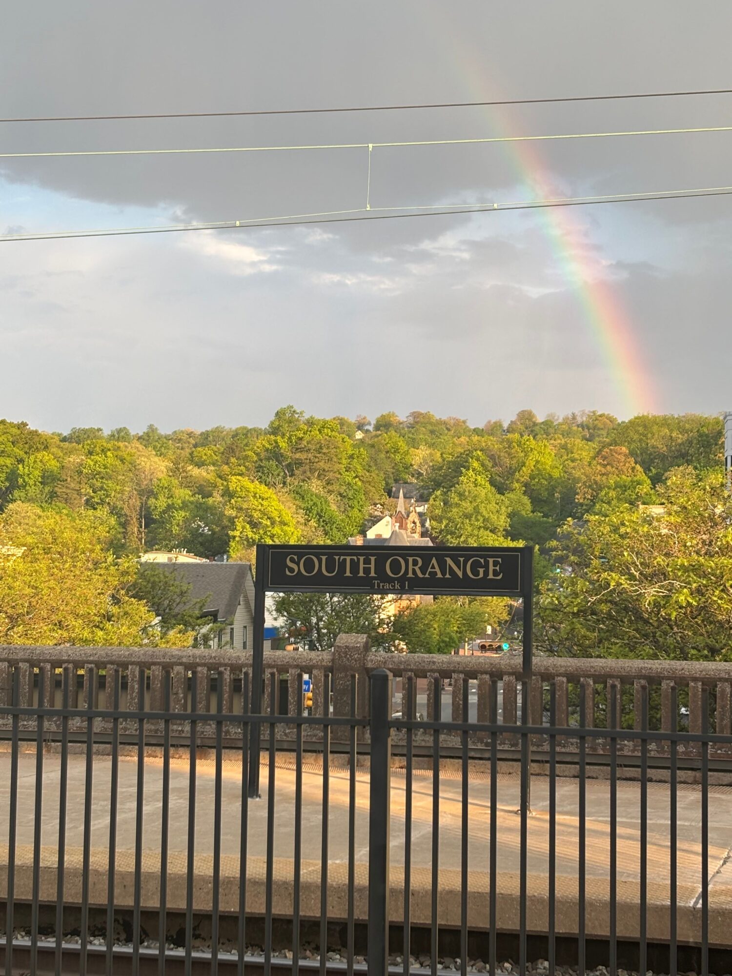 Rainbow over trees viewed from a balcony with a South Brisbane sign, cloudy sky, and sunlight.