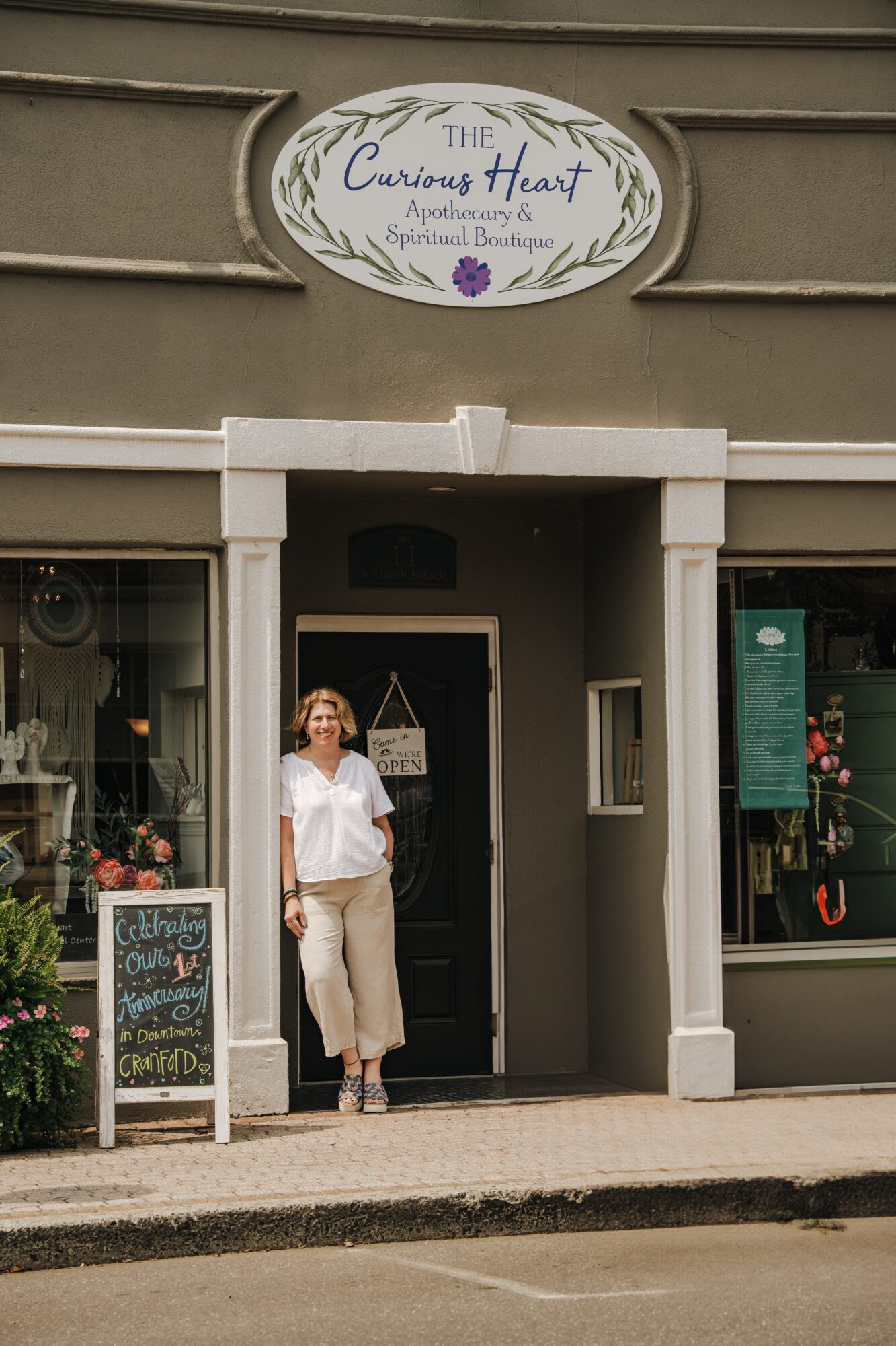 Woman standing outside storefront with sign reading 'Curious Heart' above entrance, window displays, and sidewalk in front.