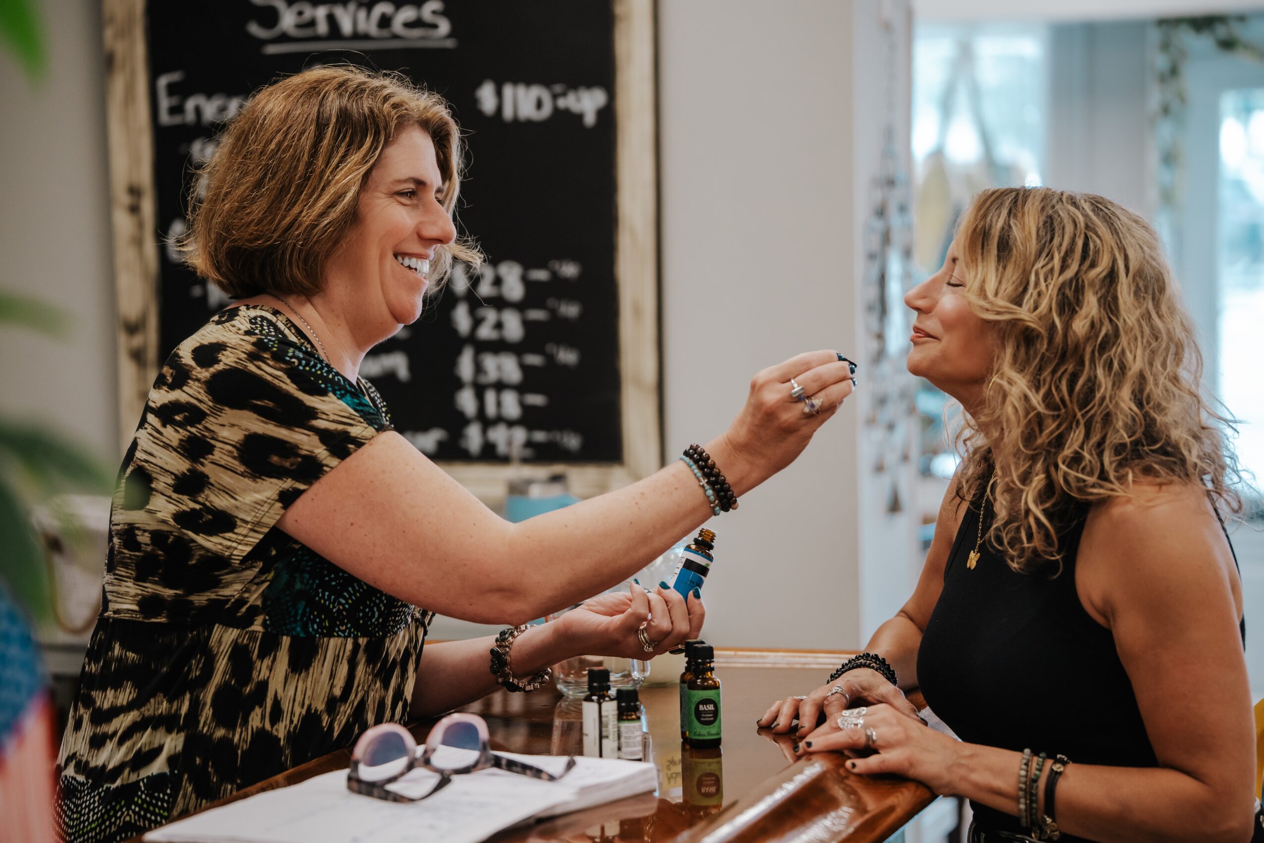 Two women smiling and talking at a table in a cafe, with a chalkboard menu in the background.
