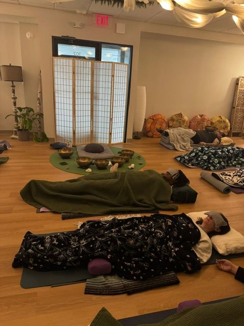 Room with yoga mats, blankets, and pillows on a wooden floor, window with blinds, and a door with an exit sign.