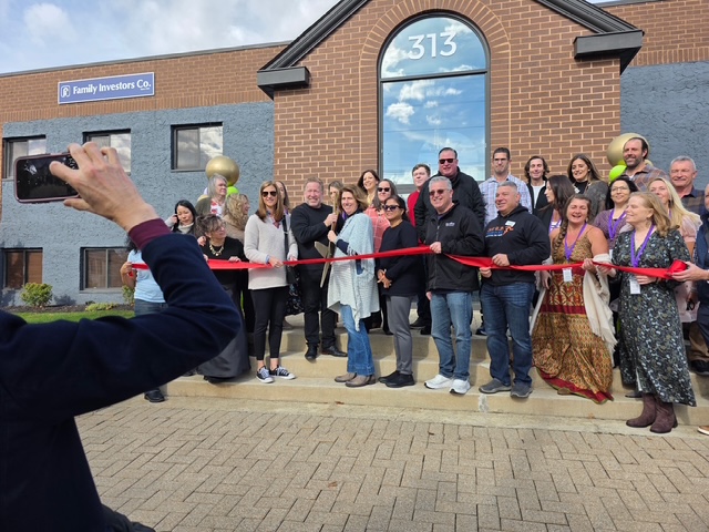 Group of people at a ribbon-cutting event outside a building with the number 313, some holding balloons, a person taking a photo.