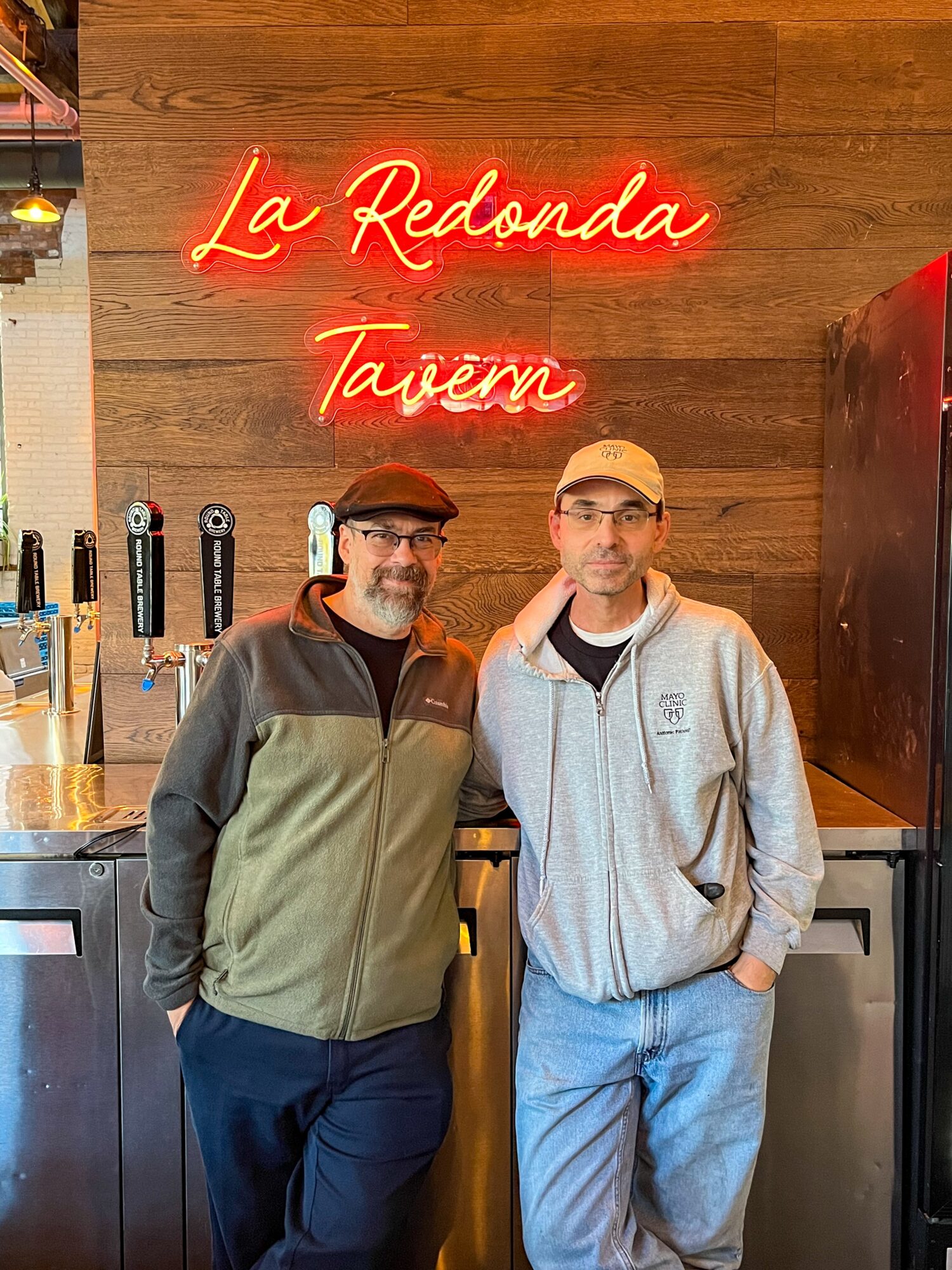 Two men stand in front of a bar with a neon sign reading 'La Redonda Tavem' on a wooden wall.
