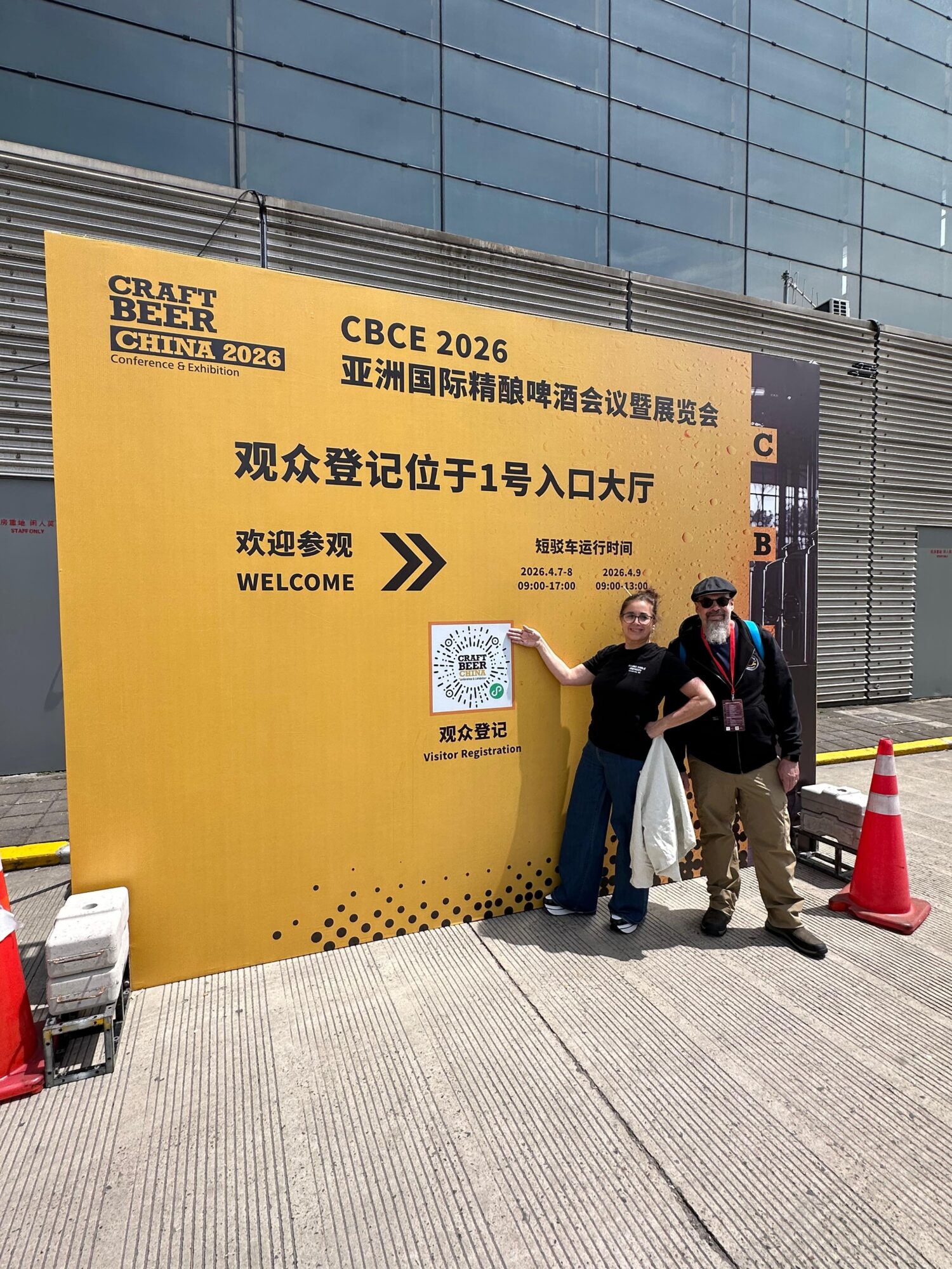 Three people standing in front of a large yellow sign with Chinese and English text, outside a modern glass building.