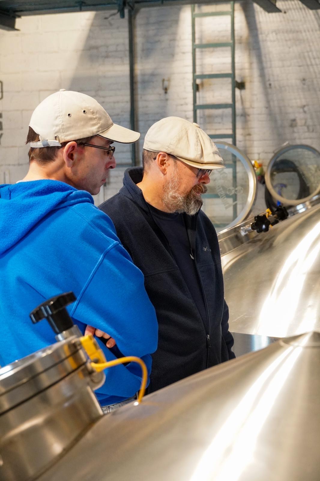 Two men wearing caps and glasses observe industrial equipment in a factory setting.