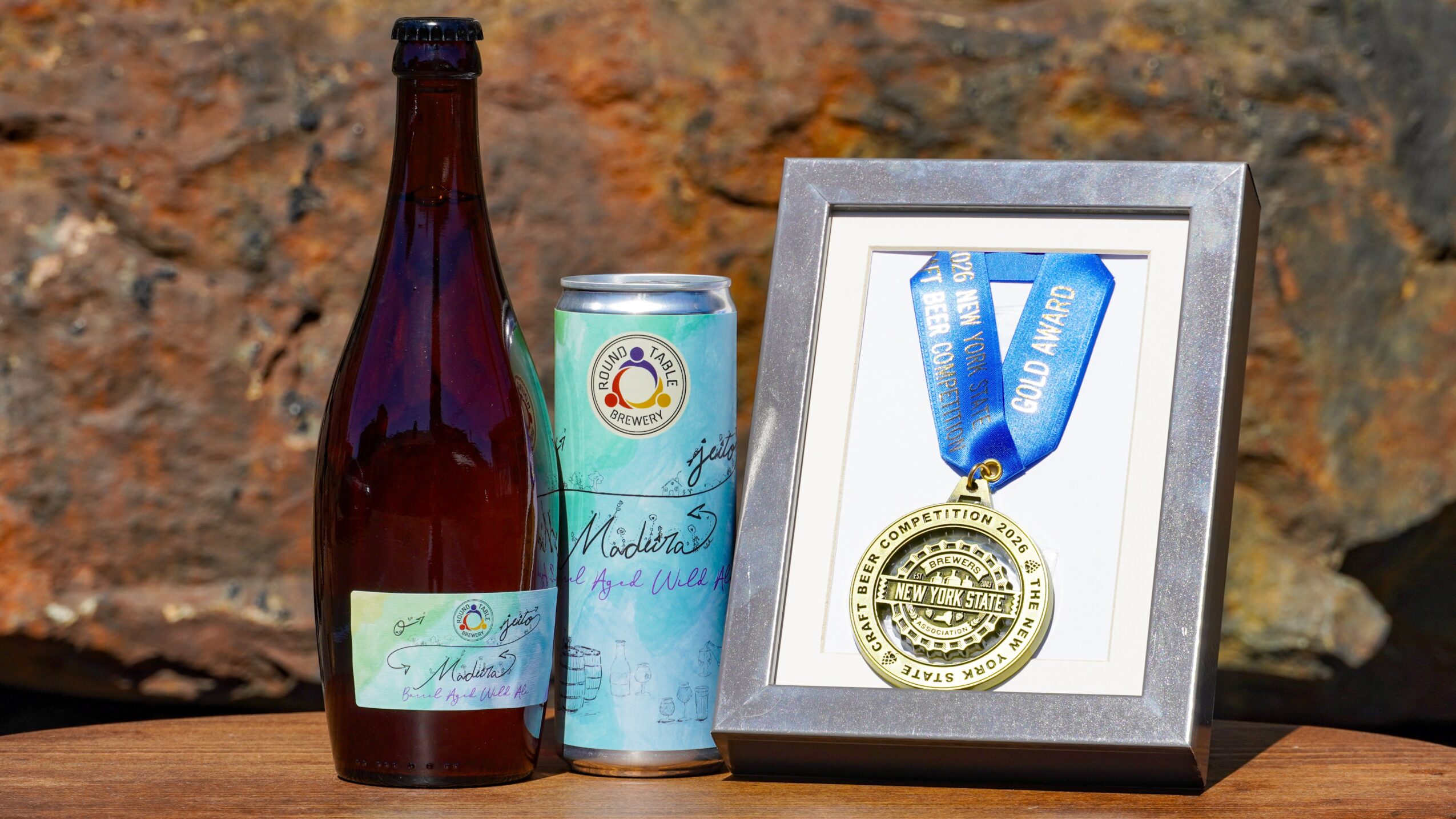 A framed gold medal with a blue ribbon, a bottle, and a can on a wooden surface against a stone background.