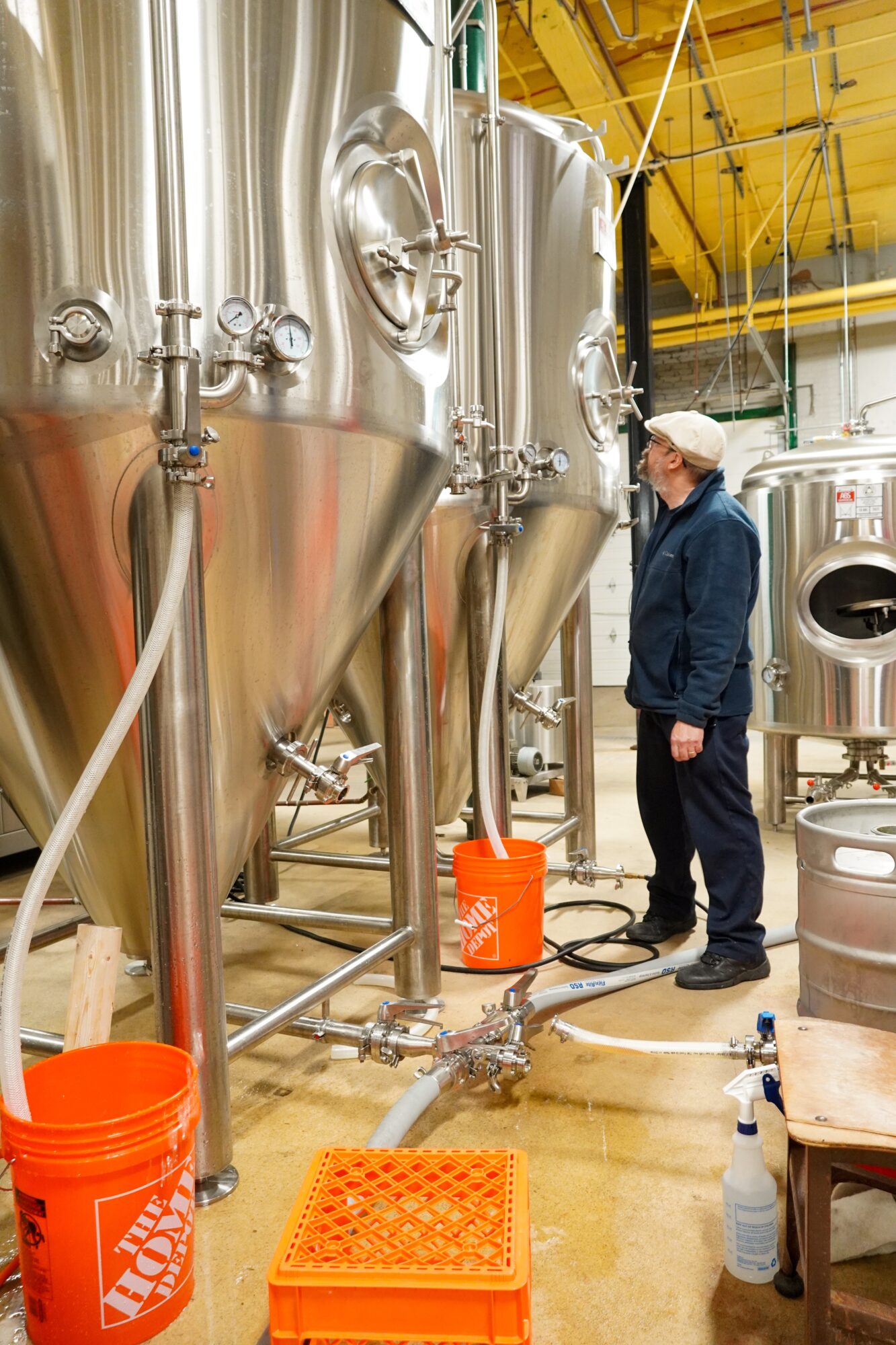 Person in a brewery or industrial facility with large stainless steel tanks, wearing a cap and dark clothing, standing near equipment.