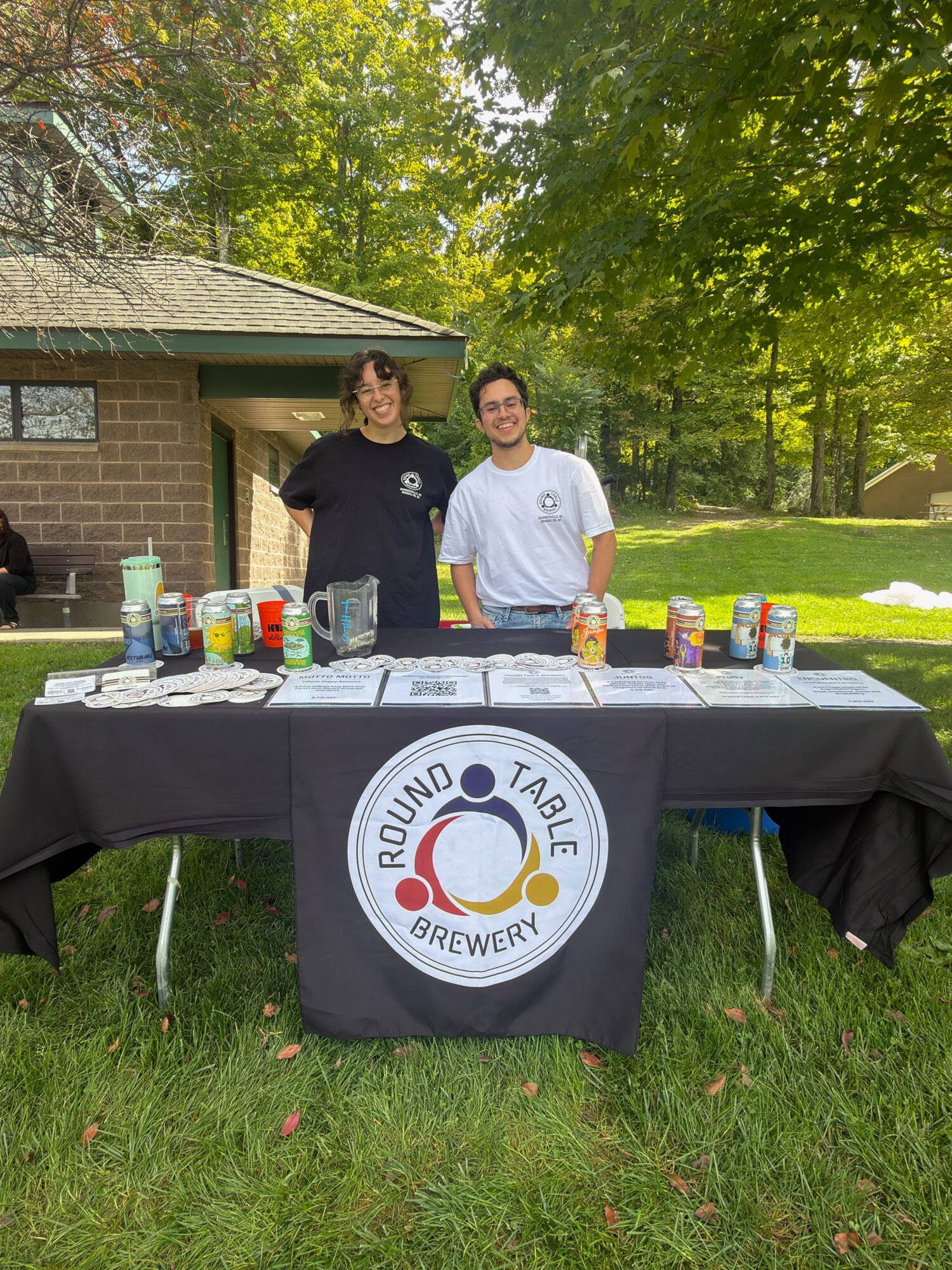 Two people standing behind a table with drinks outdoors, table has a round logo with a ring and text, surrounded by trees.