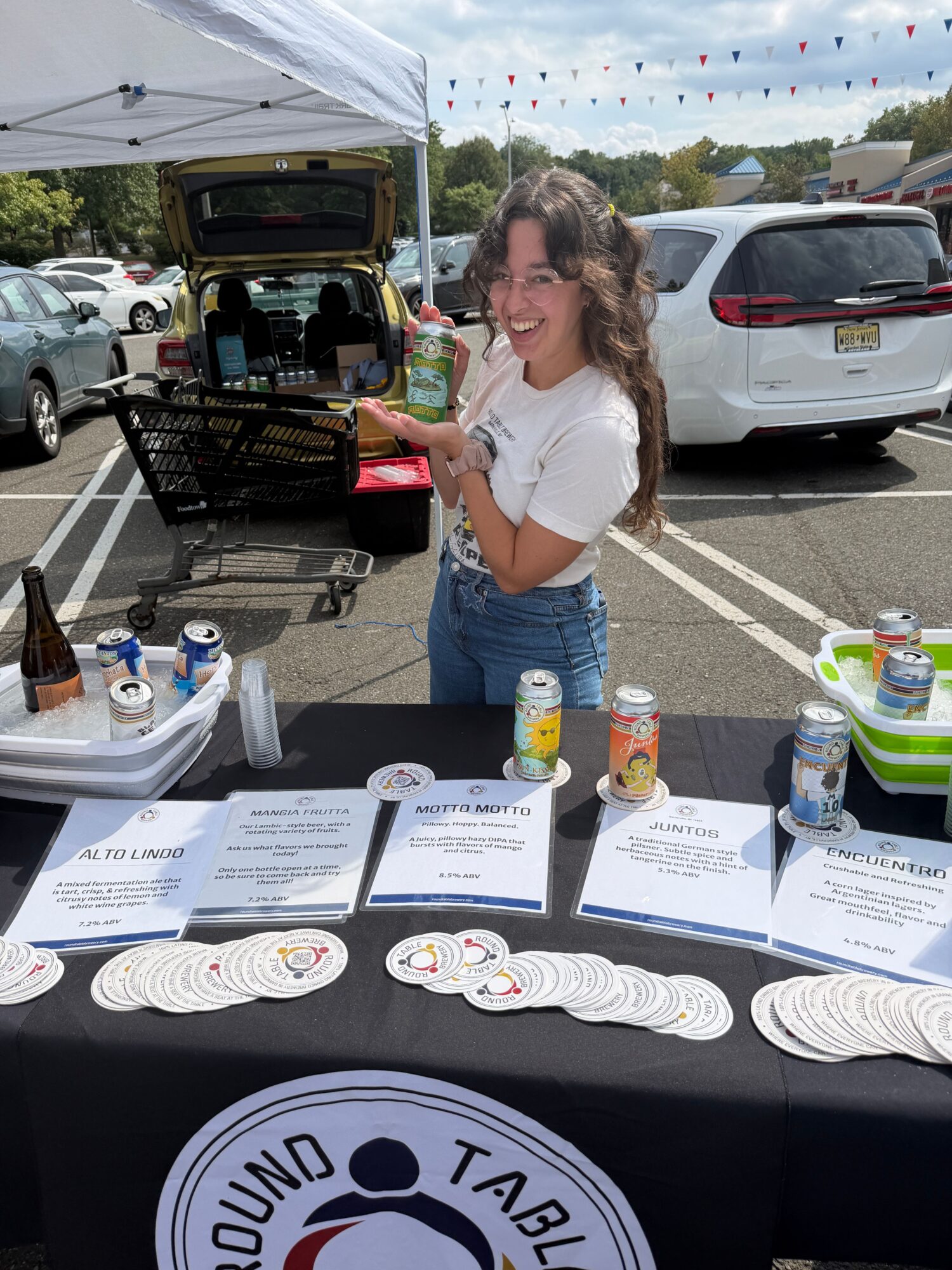 Woman at outdoor table with food, drinks, and informational materials, with cars and trees in background.