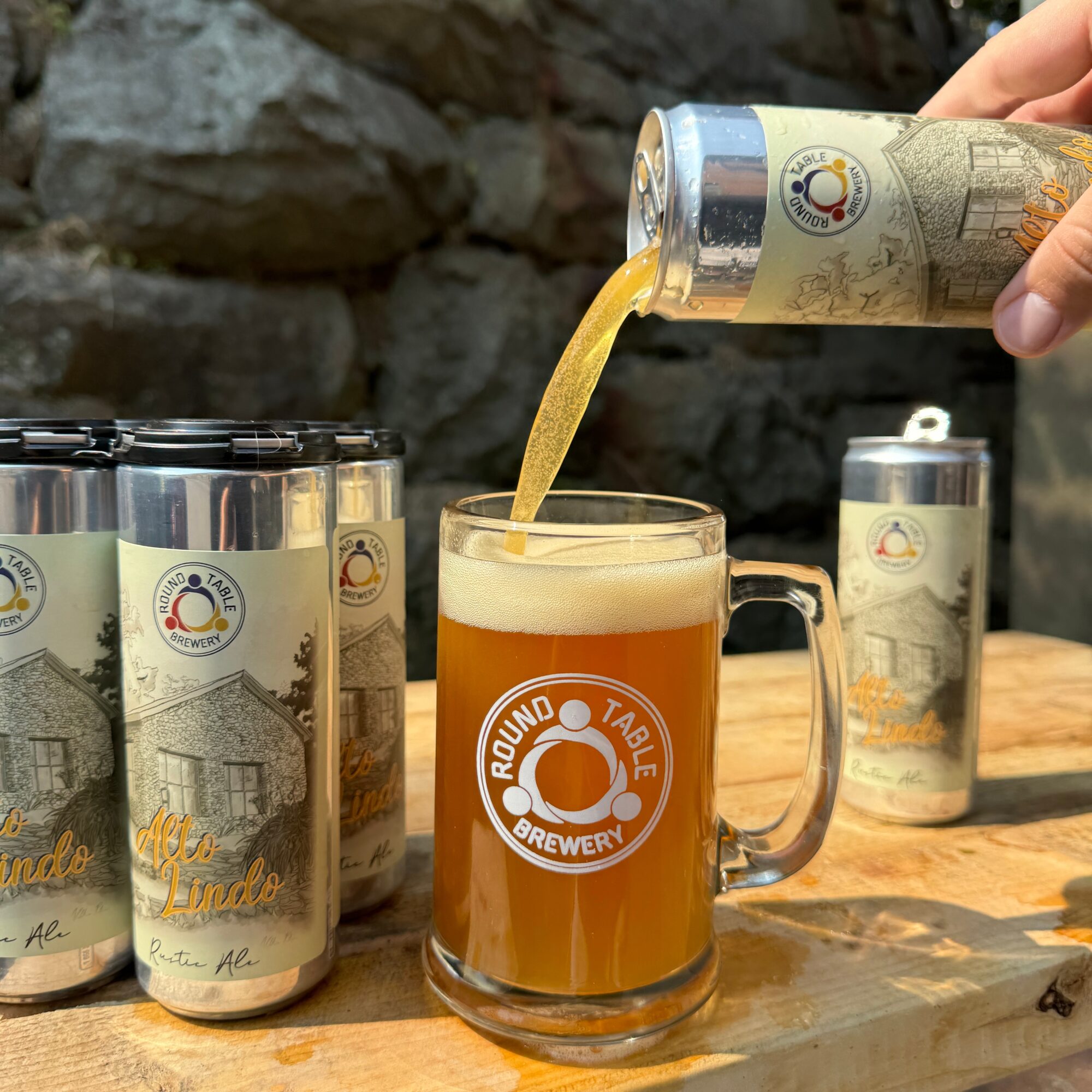 Person pouring beer from a can into a glass mug with a brewery logo, on a wooden table with cans nearby.