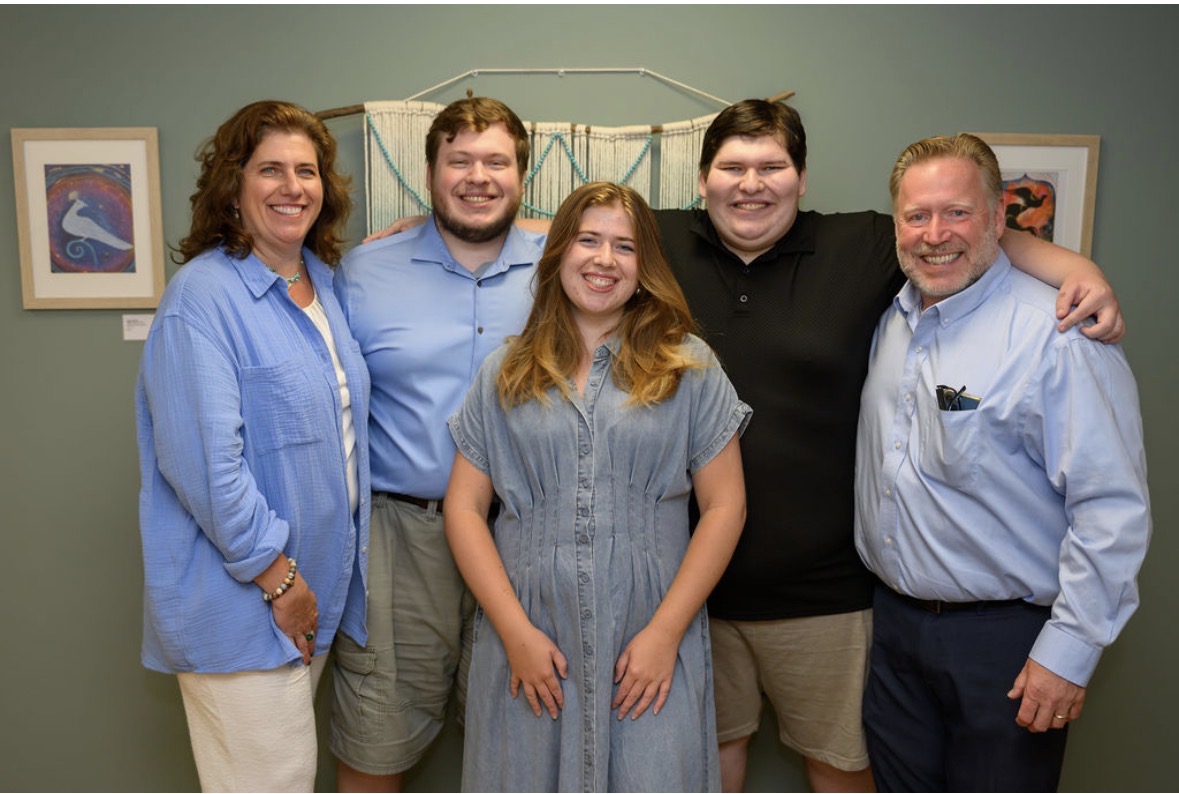 Five people standing together, smiling, in a room with framed pictures on the wall.