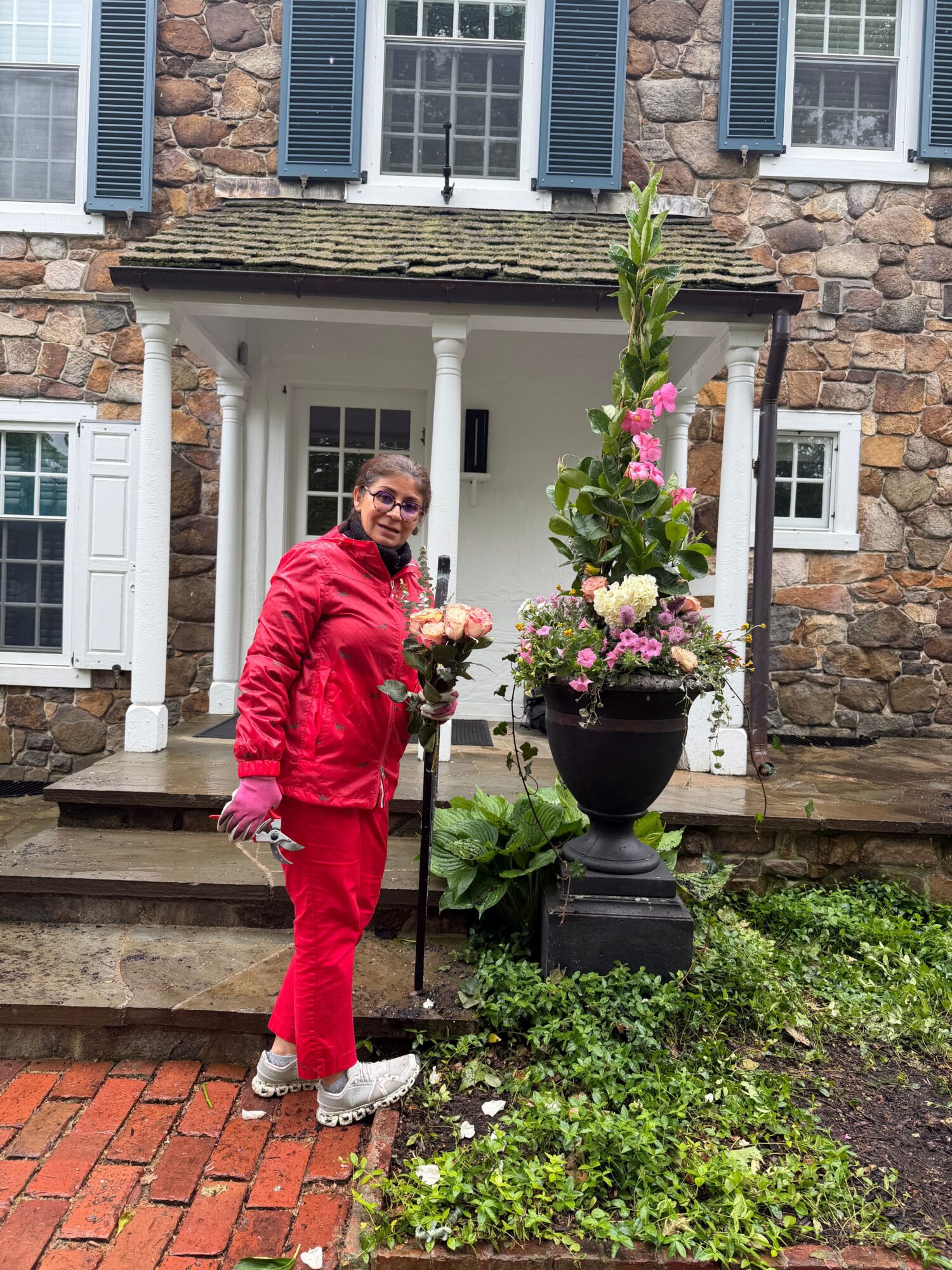 Person in red outfit holding a bouquet of flowers, standing outside near a stone wall and garden, smiling.
