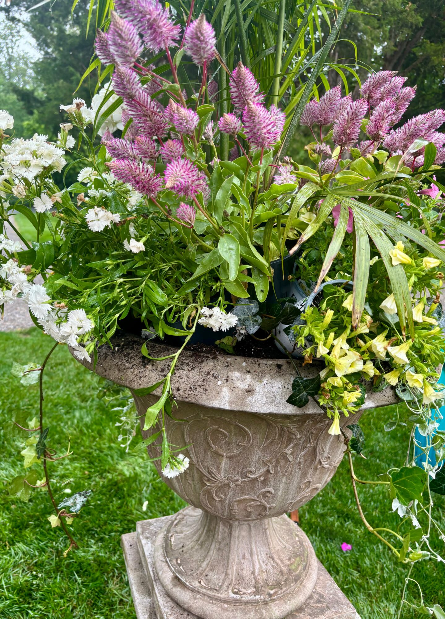 Flower arrangement in a decorative stone planter with purple, white, and yellow flowers, outdoors with green grass and trees.