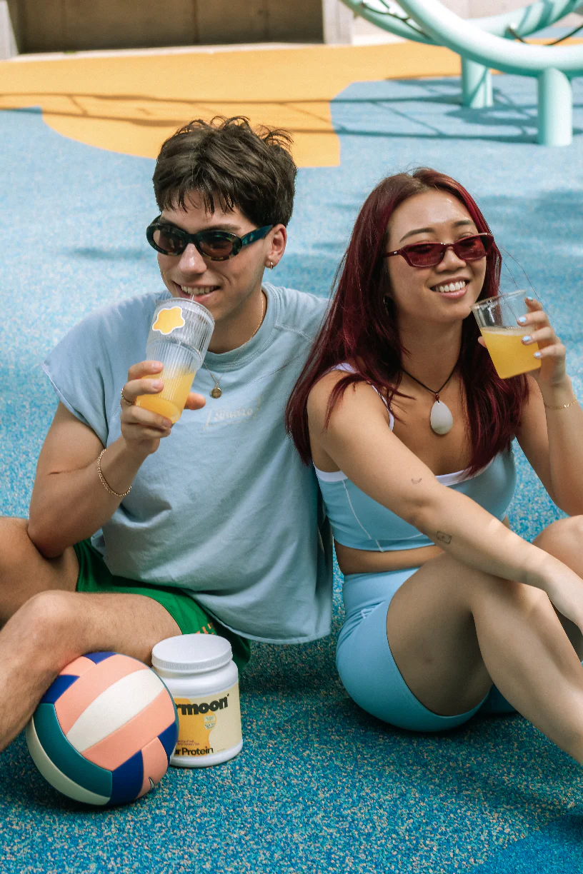Two people sitting on a poolside, smiling, holding drinks, with a volleyball and sunscreen nearby.