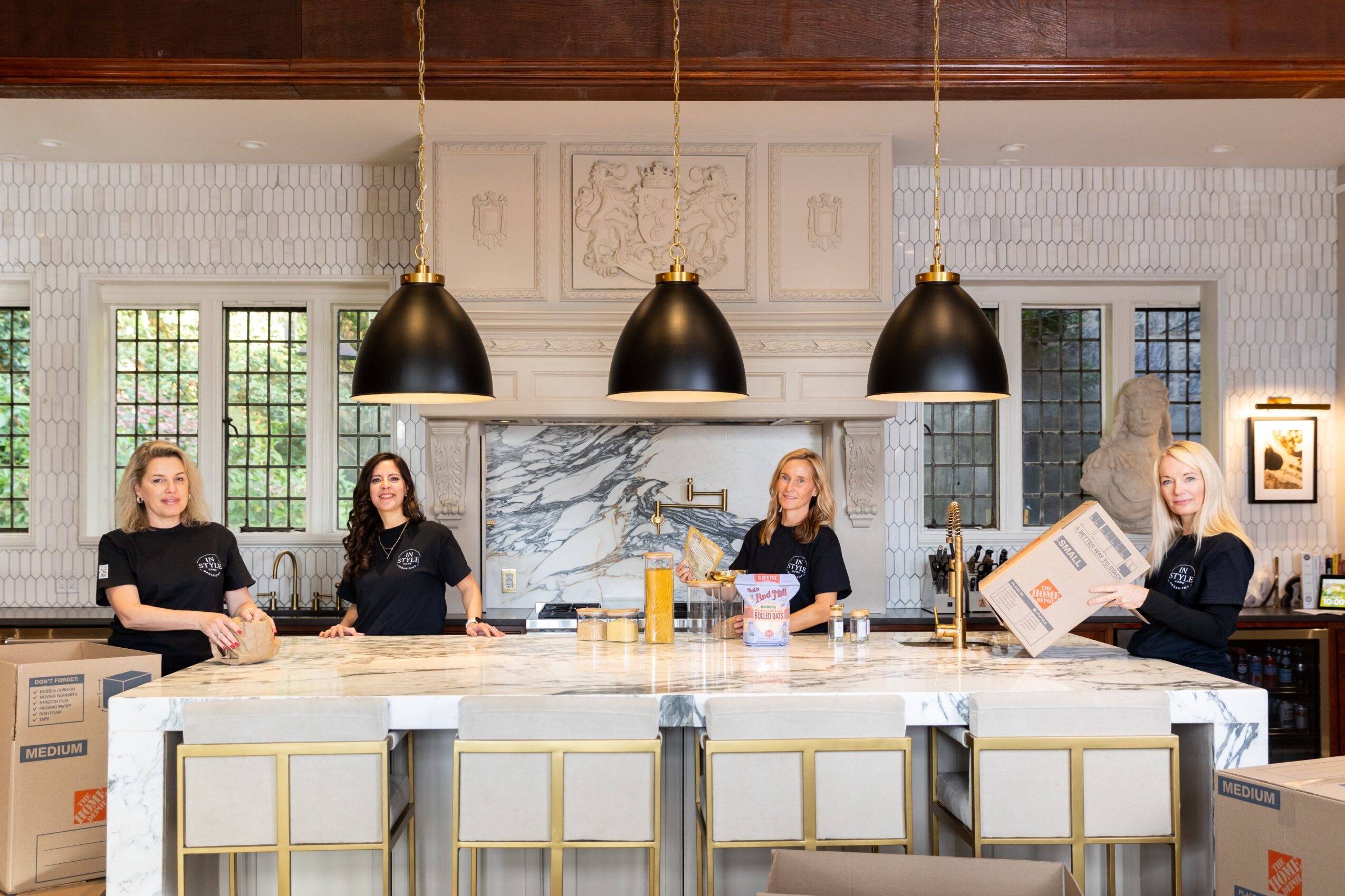 Five women stand behind a kitchen island with white cabinets, black pendant lights, and a marble backsplash, in a bright room.