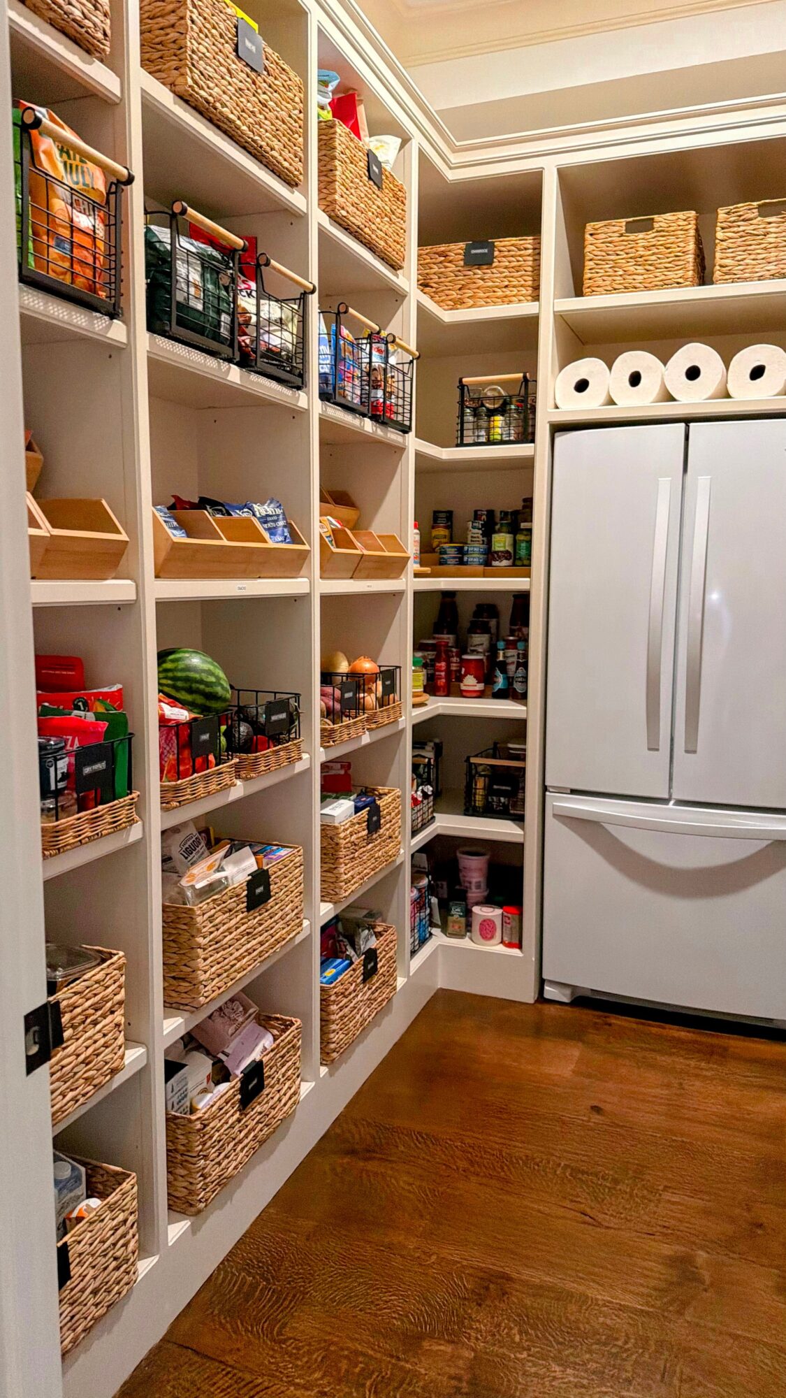 Pantry with shelves filled with baskets, food items, and a refrigerator, wooden floor, and ceiling lighting.