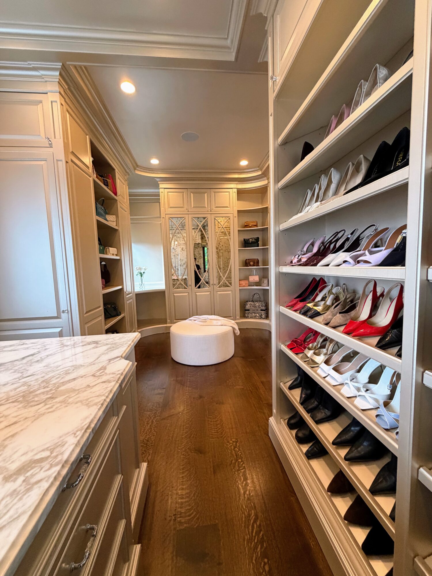 Walk-in closet with shelves of shoes, a marble countertop, and a white ottoman in the center, with built-in cabinets and hardwood floors.