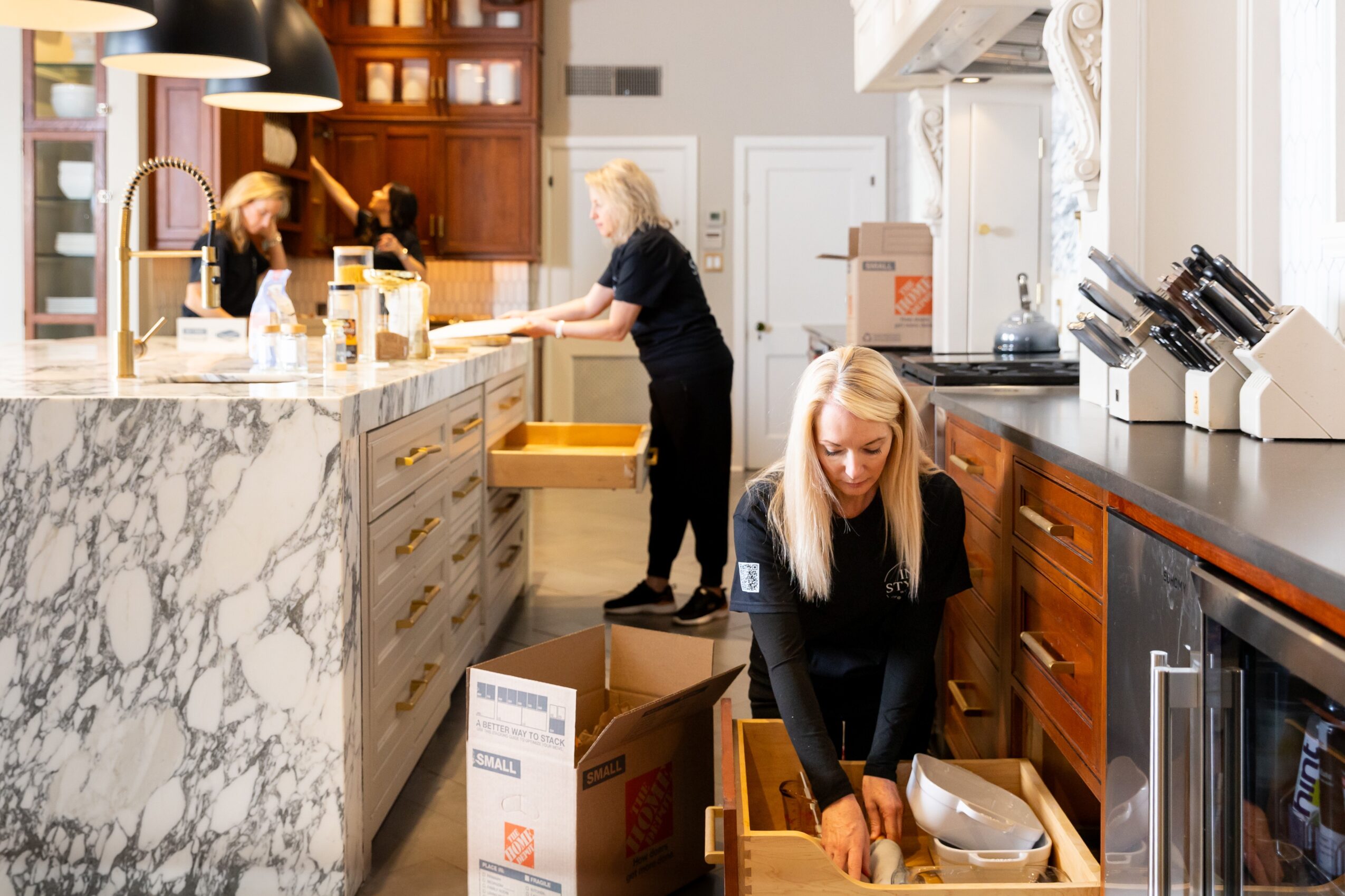 Three women in a kitchen with marble countertops and wooden cabinets, one packing items into a box, another at the counter, and a third in the background.