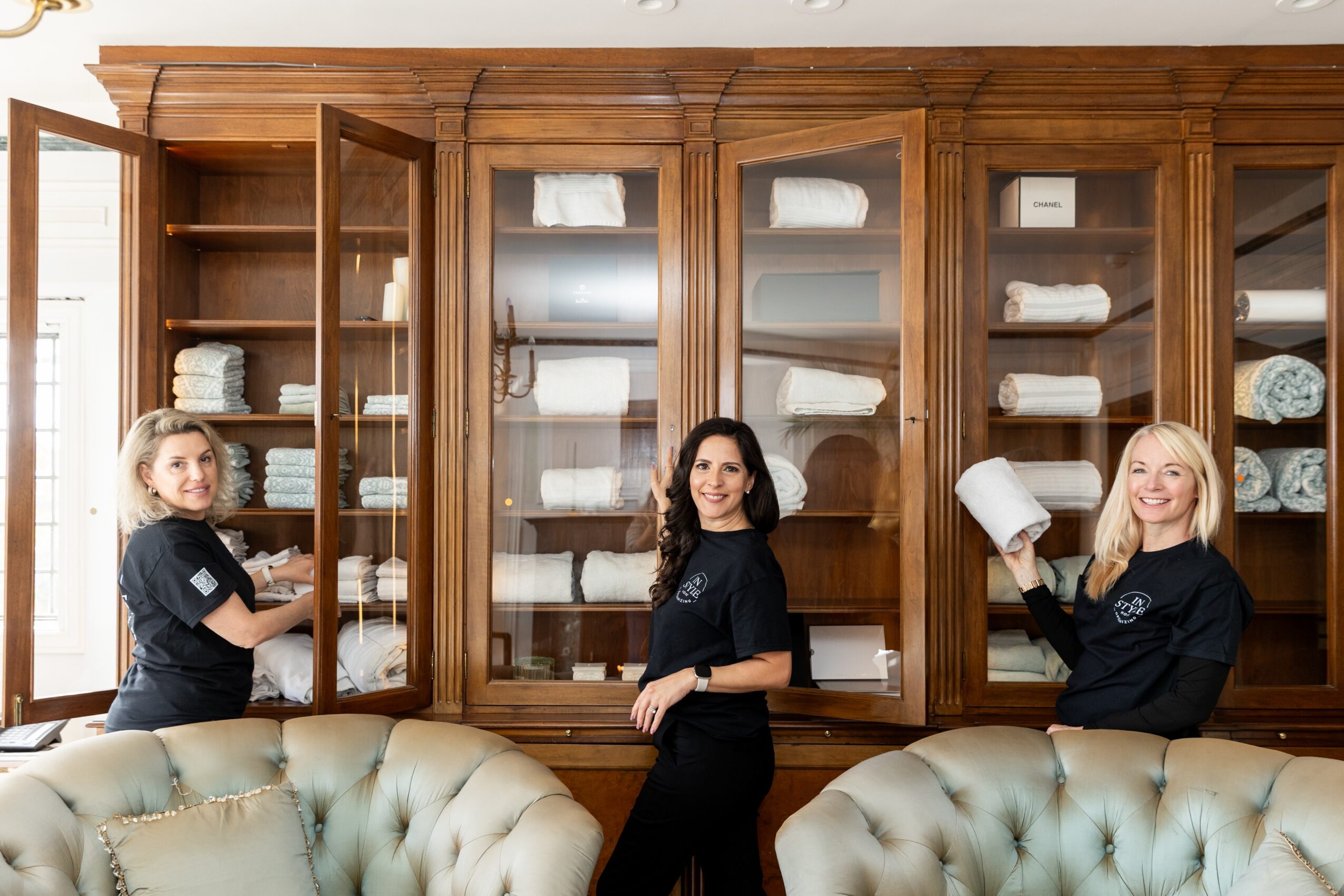 Three women in black uniforms stand in front of a large wooden cabinet filled with neatly folded towels, smiling.