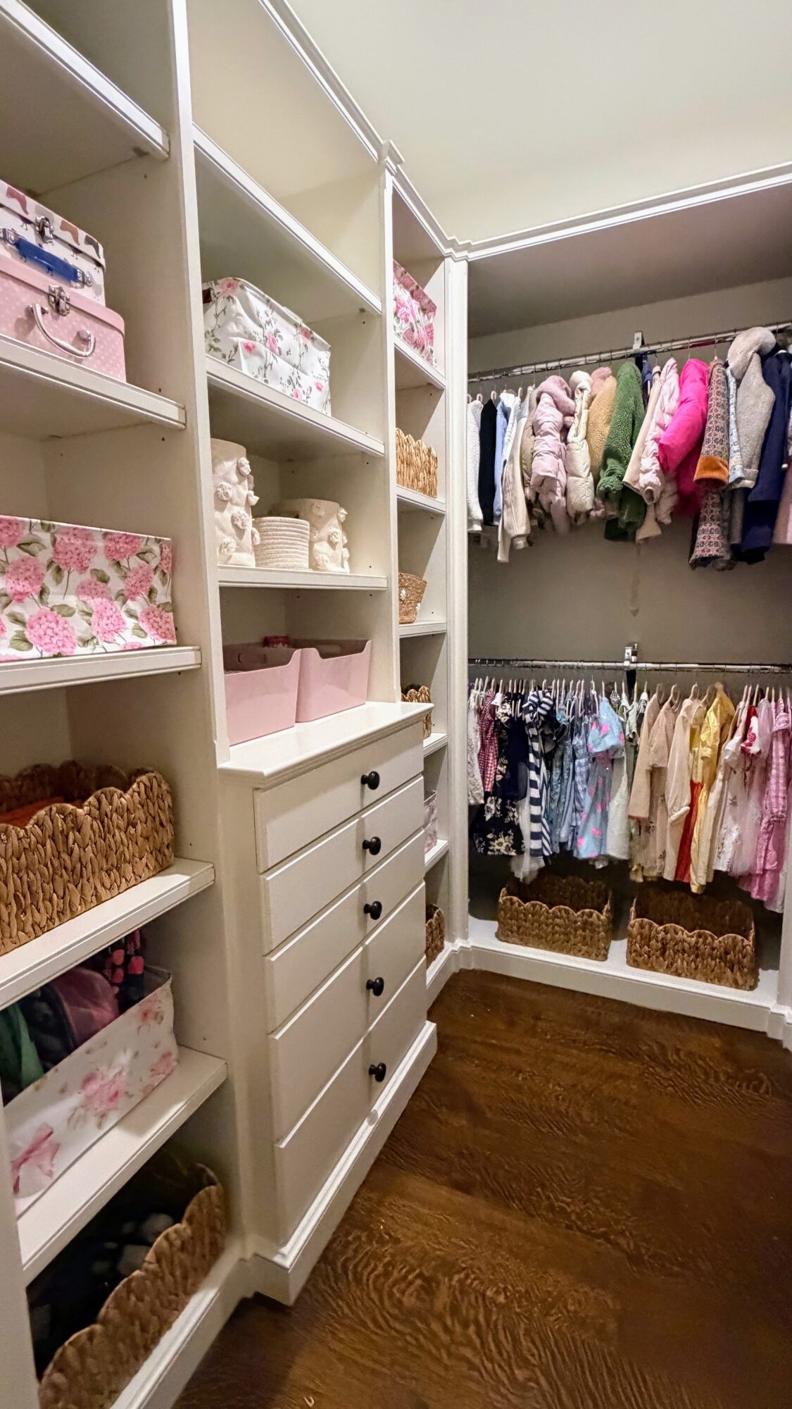 Closet with shelves and drawers filled with folded clothes, baskets, and hanging garments, wooden floor, and ceiling light.