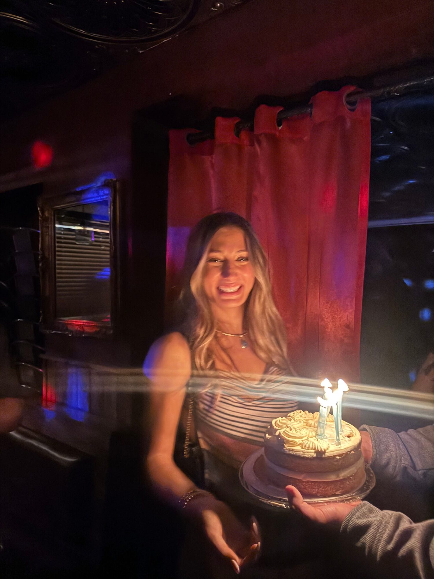 Woman celebrating birthday with cake and lit candles, red curtains in background, dim lighting.