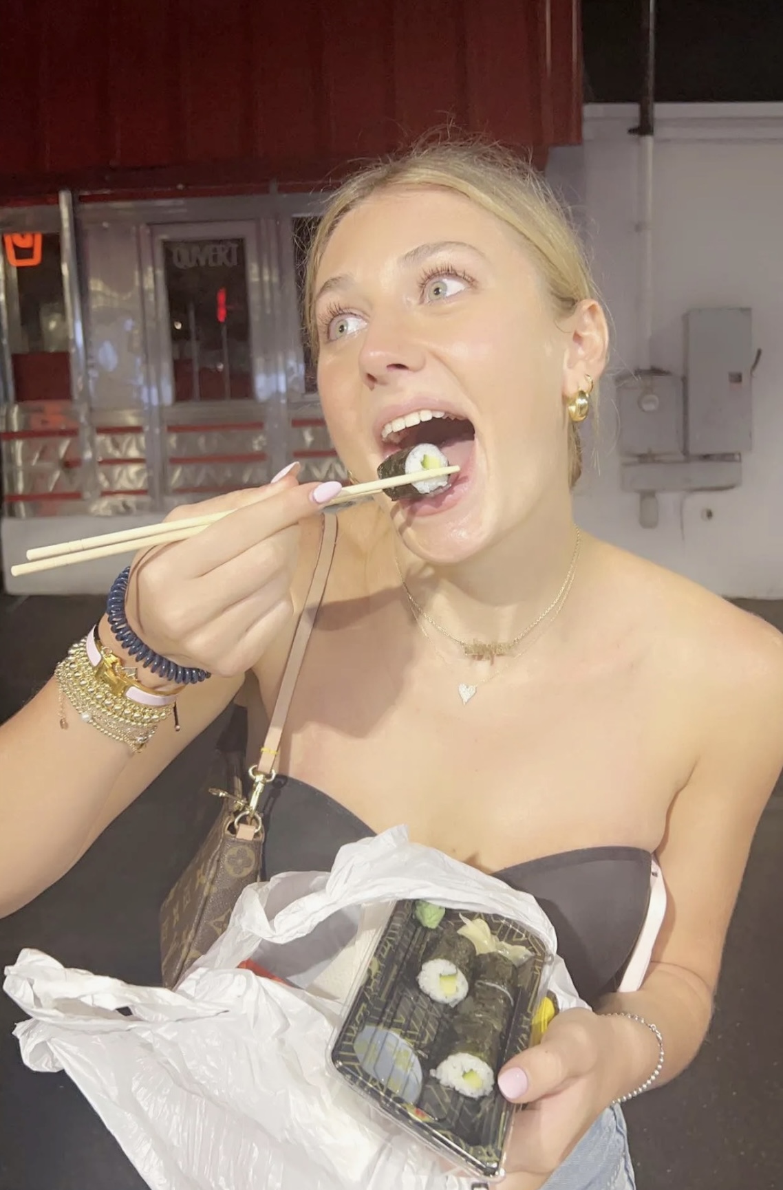 Woman eating sushi with chopsticks, holding a container of sushi, in a restaurant setting.