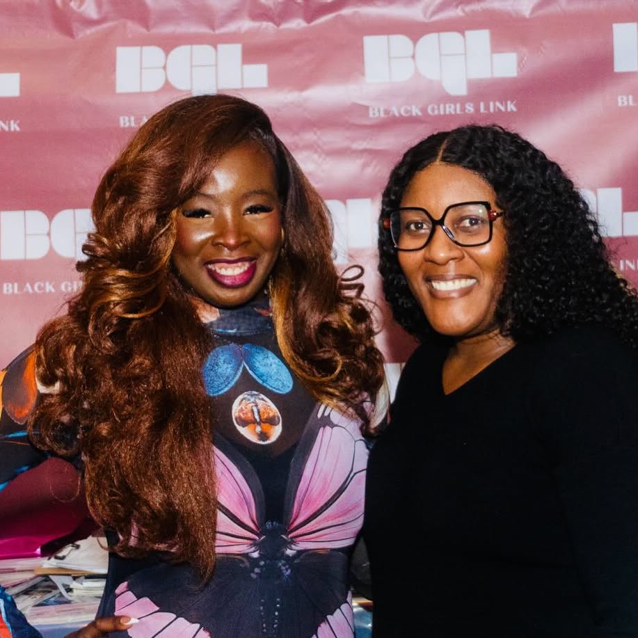 Two women smiling in front of a pink backdrop with white text, one with long wavy hair and glasses, the other with curly hair.