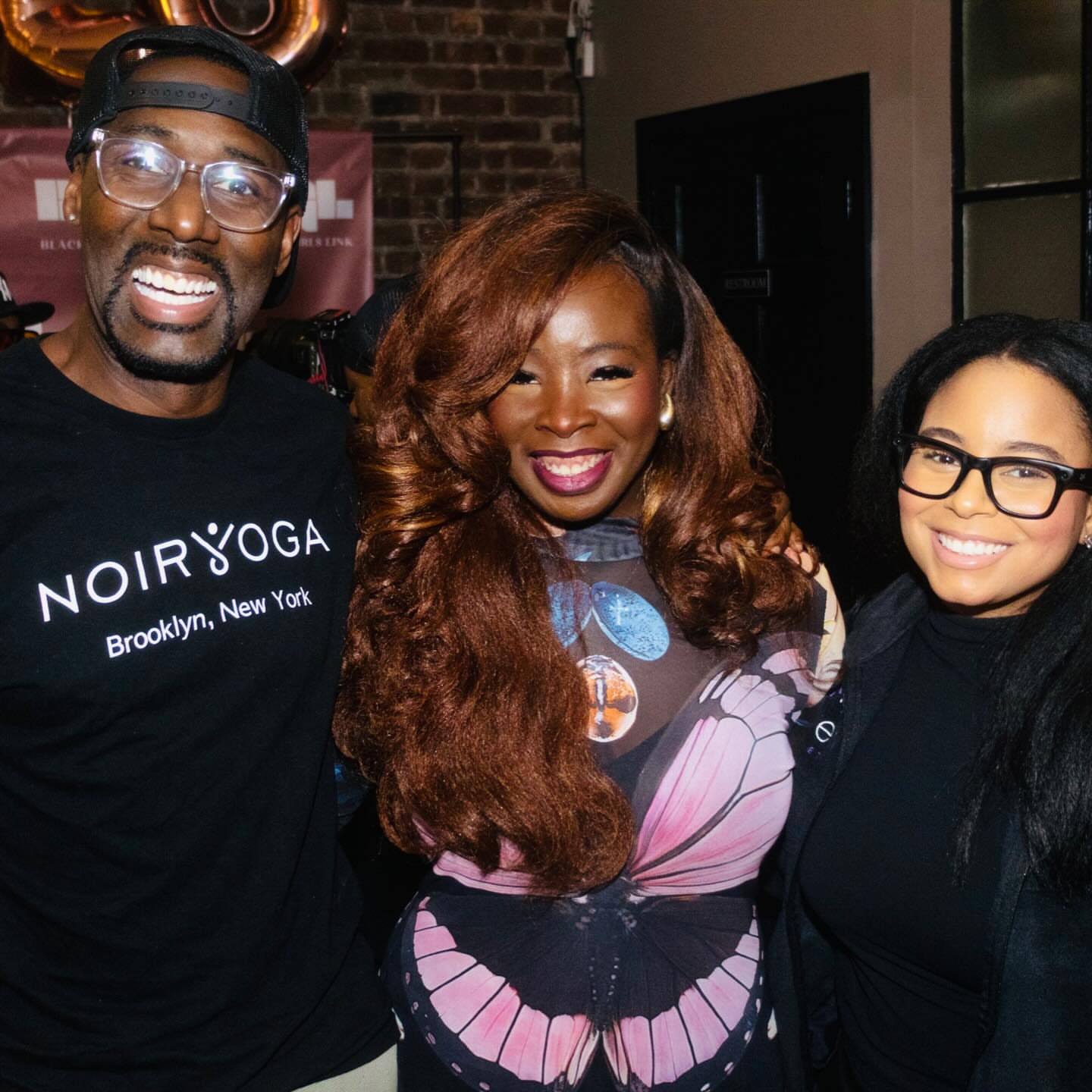 Three people smiling indoors, two women and one man, with brick wall background, casual attire, and glasses.