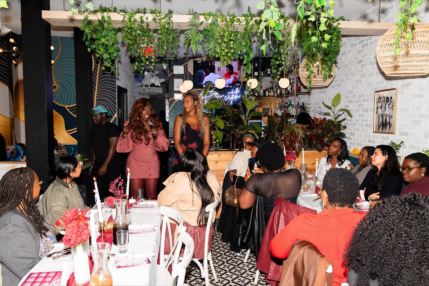 People gathered around tables with flowers and drinks in a decorated indoor space with hanging plants and artwork.