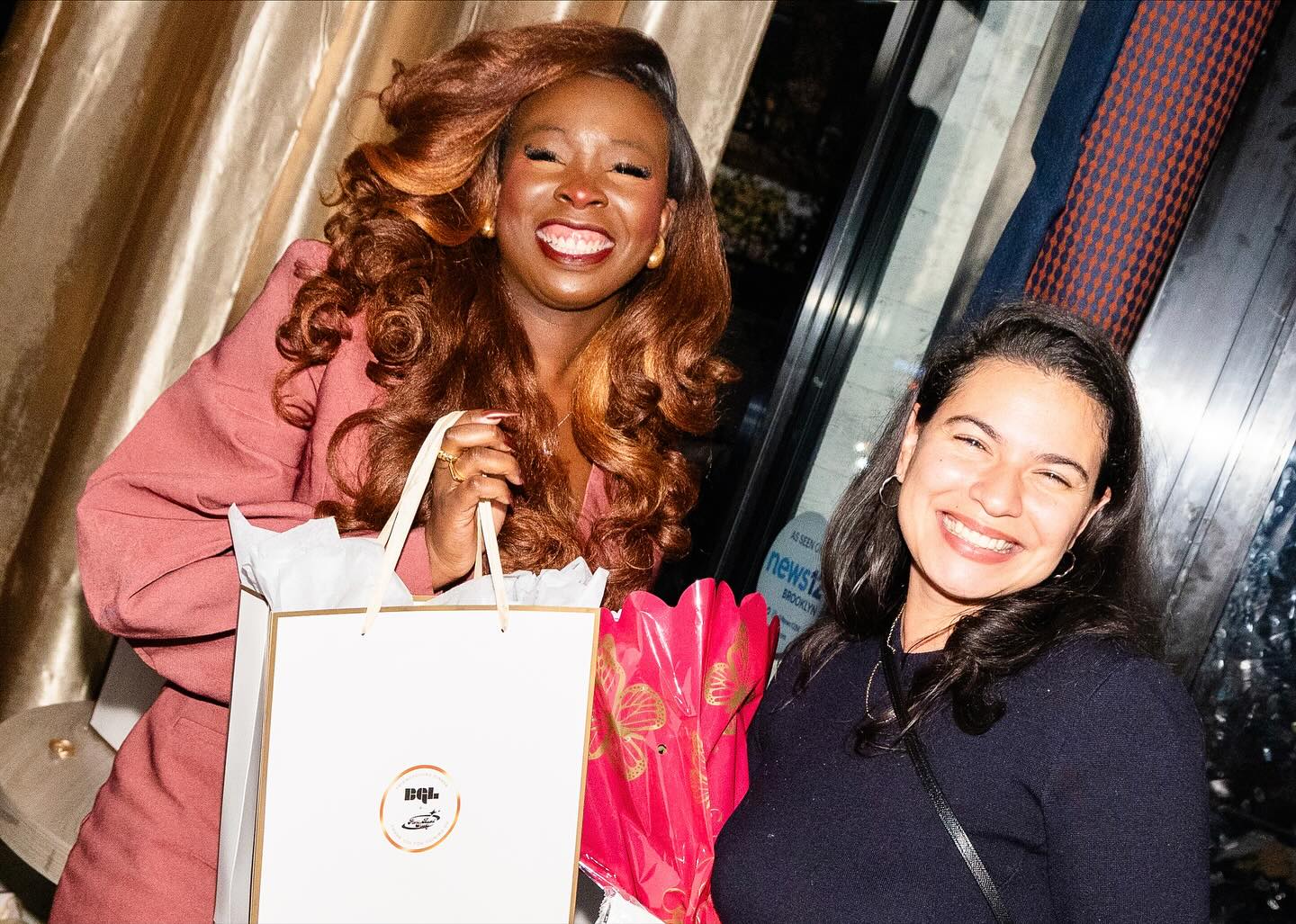 Two women smiling, one holding a shopping bag, in an indoor setting with curtains and windows.