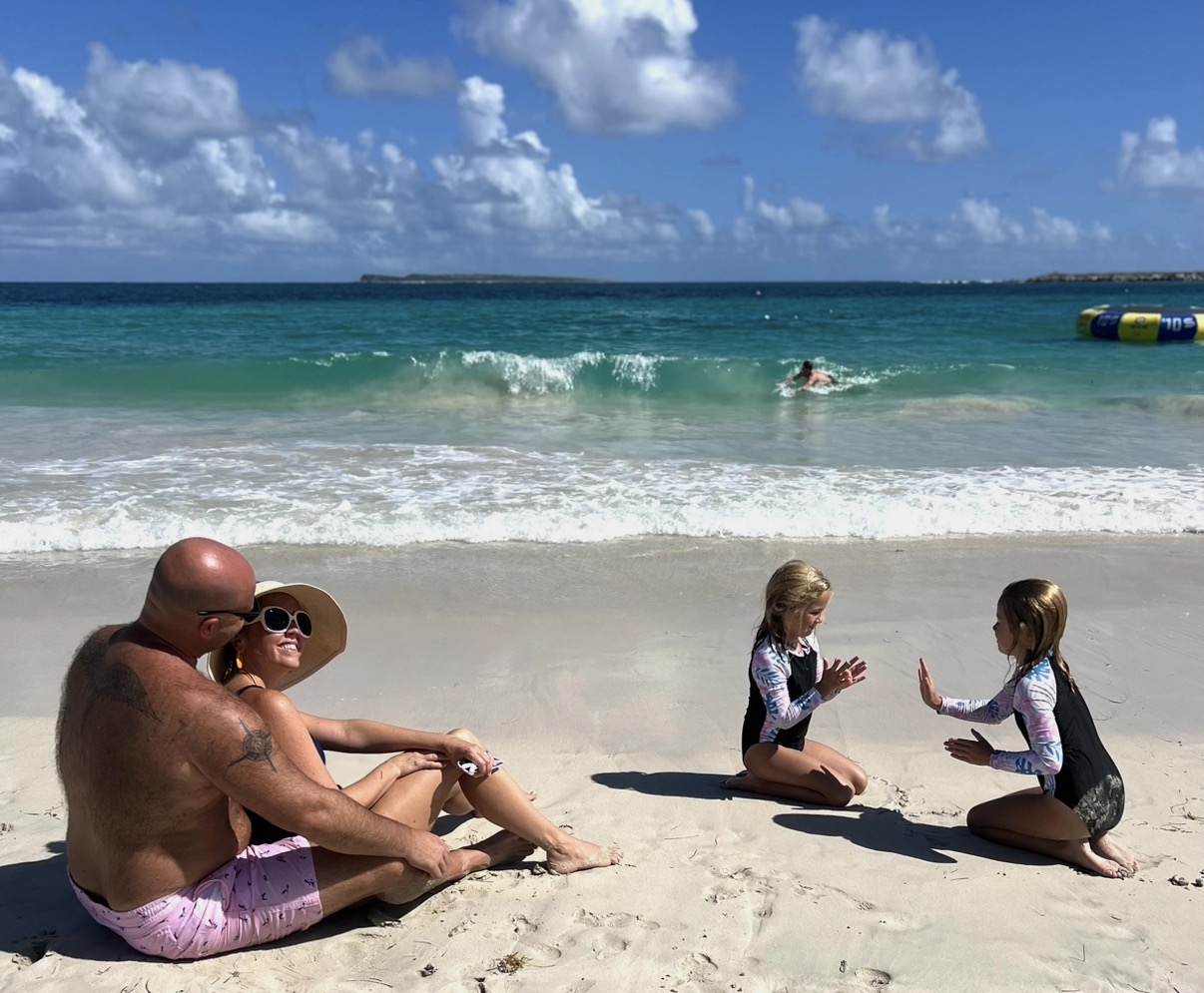Two children sit on the beach facing each other, talking, while a man and a woman sit nearby. The ocean and sky are in the background.