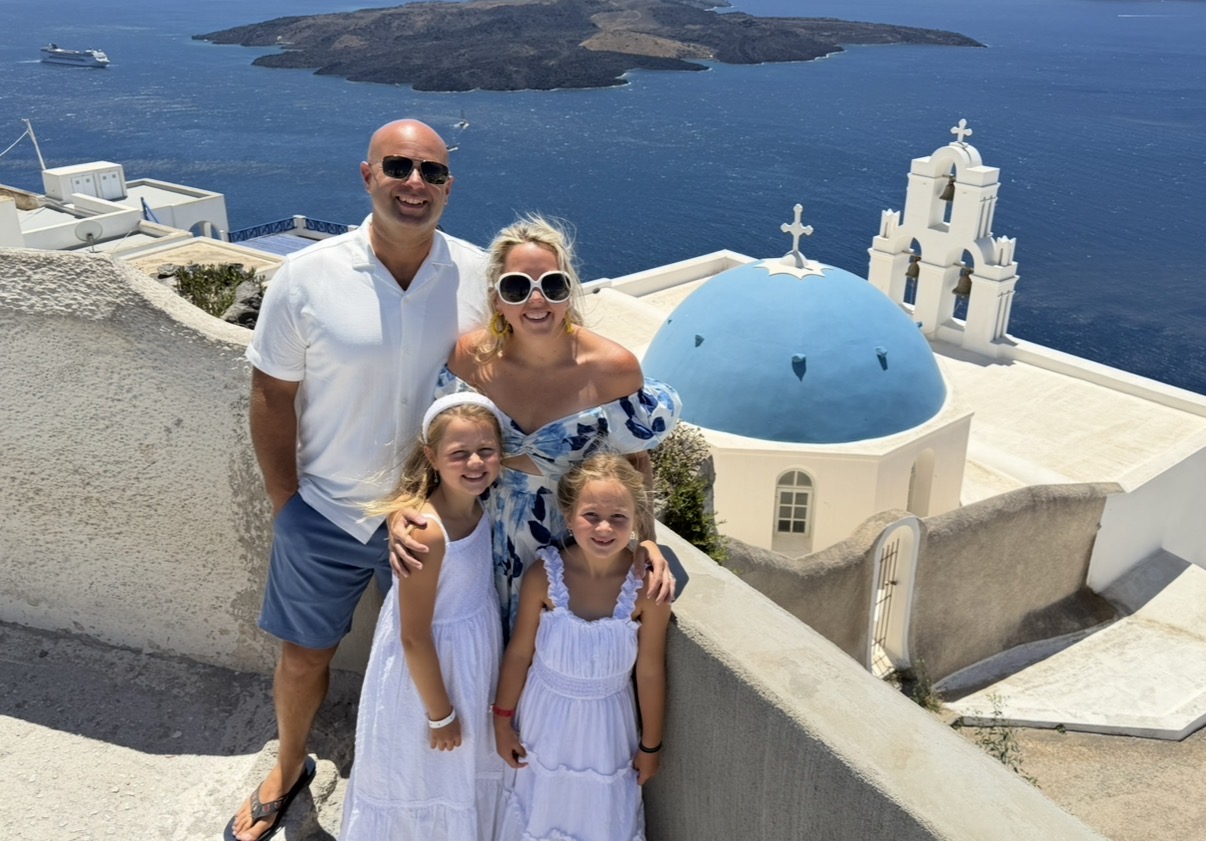 A family of four stands on a hillside with a blue-domed church and ocean in background, smiling at the camera.
