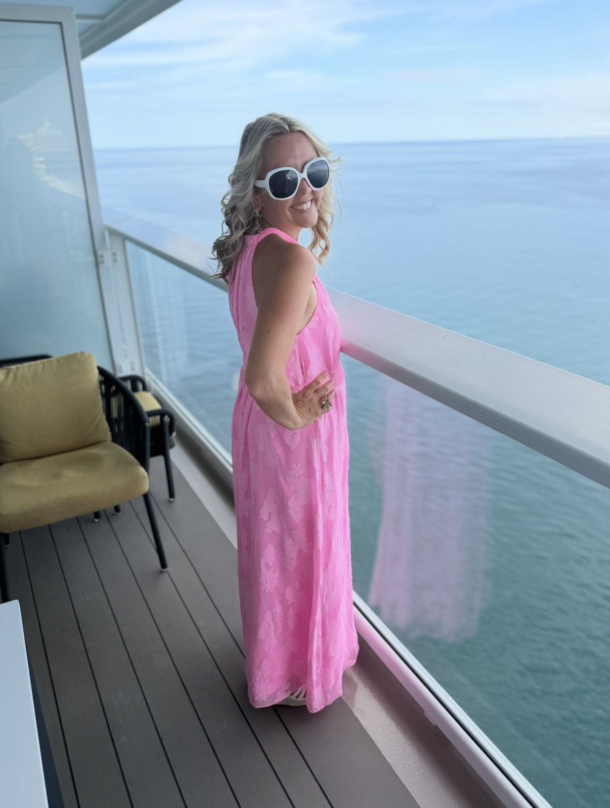 Woman in pink dress and sunglasses standing on balcony overlooking ocean, with chair nearby.